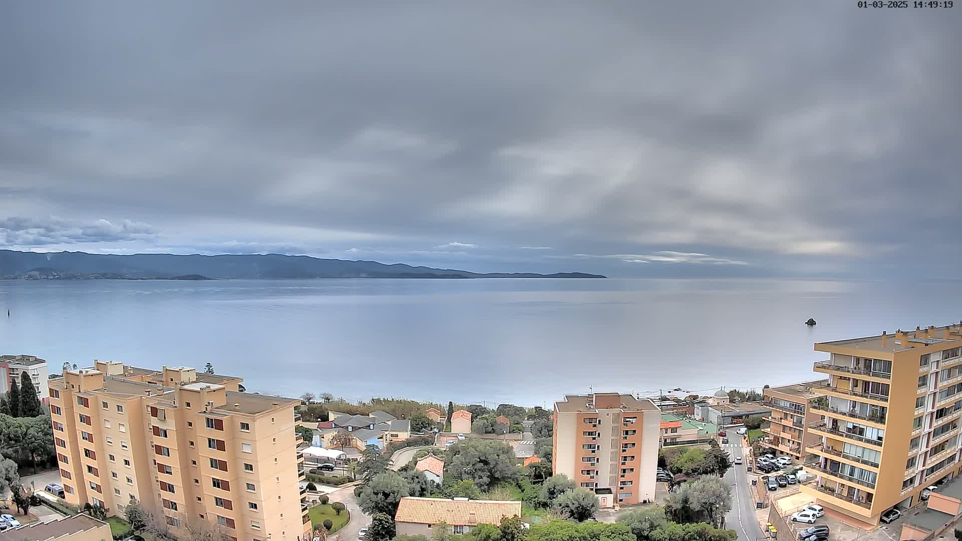 An overcast sky hangs over a calm sea, with buildings and coastal vegetation visible in the foreground.