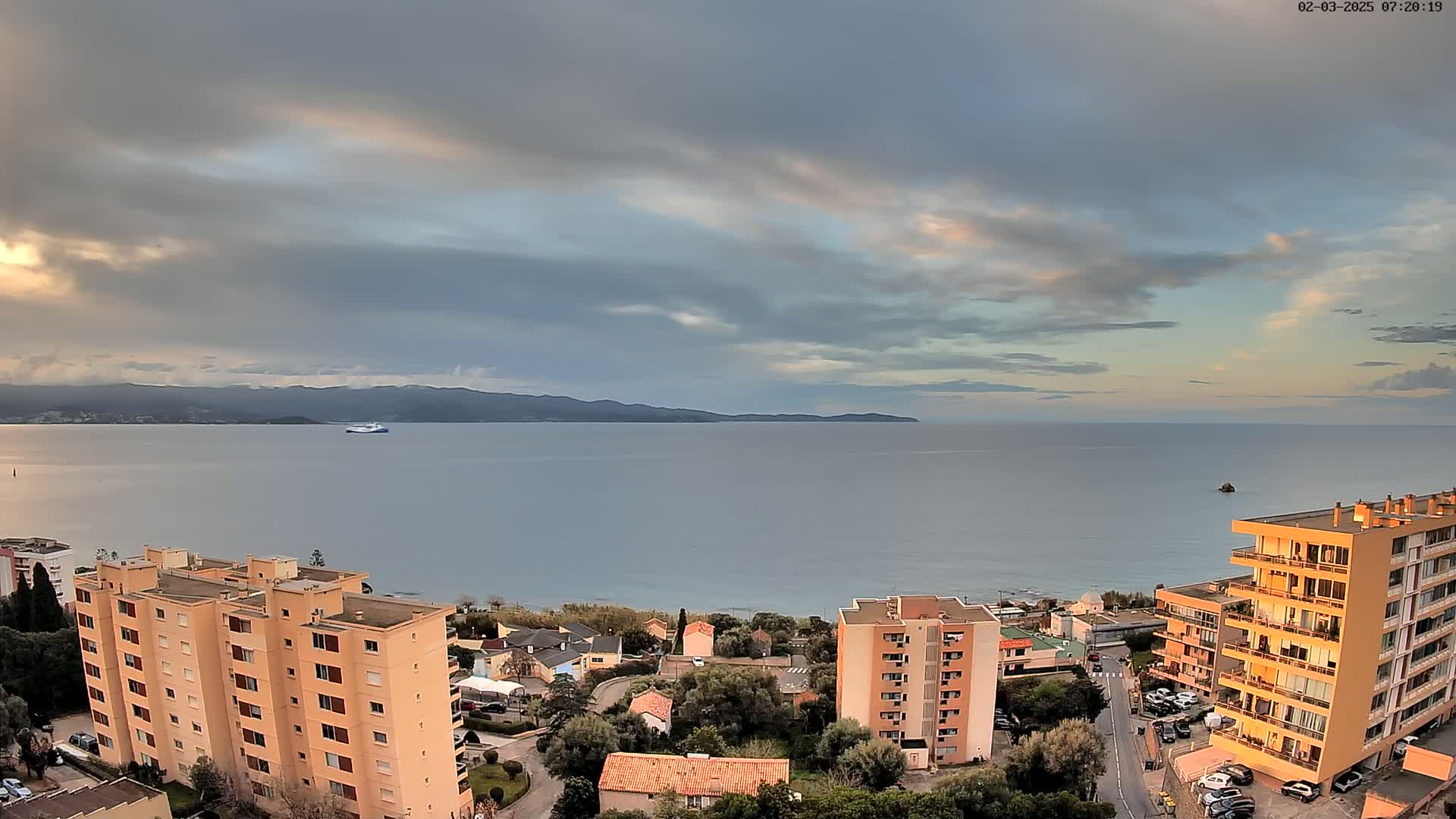 An overcast sky with hints of sunrise or sunset over a calm sea, featuring a large vessel in the distance and several multi-story buildings near a coastline.