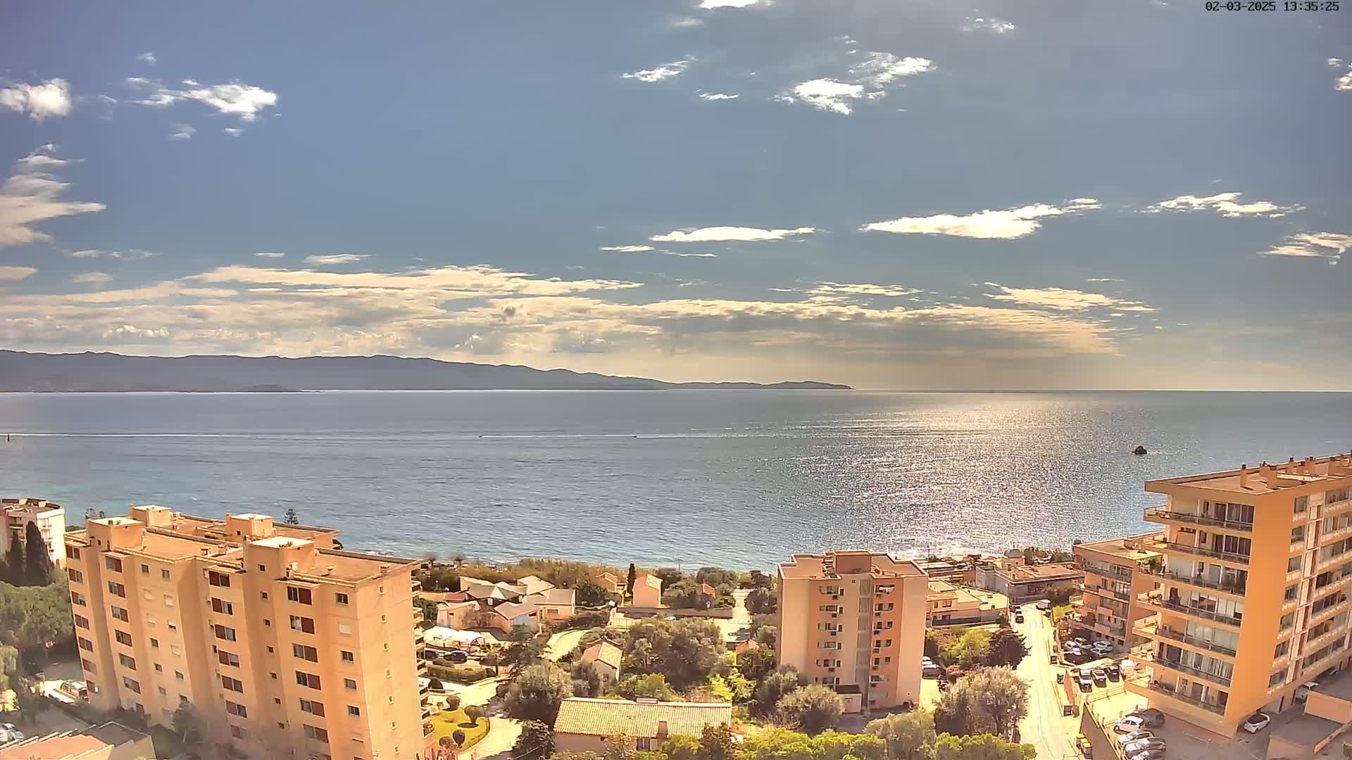 A partly cloudy day overlooks a calm sea with distant mountains, residential buildings, and a street below.