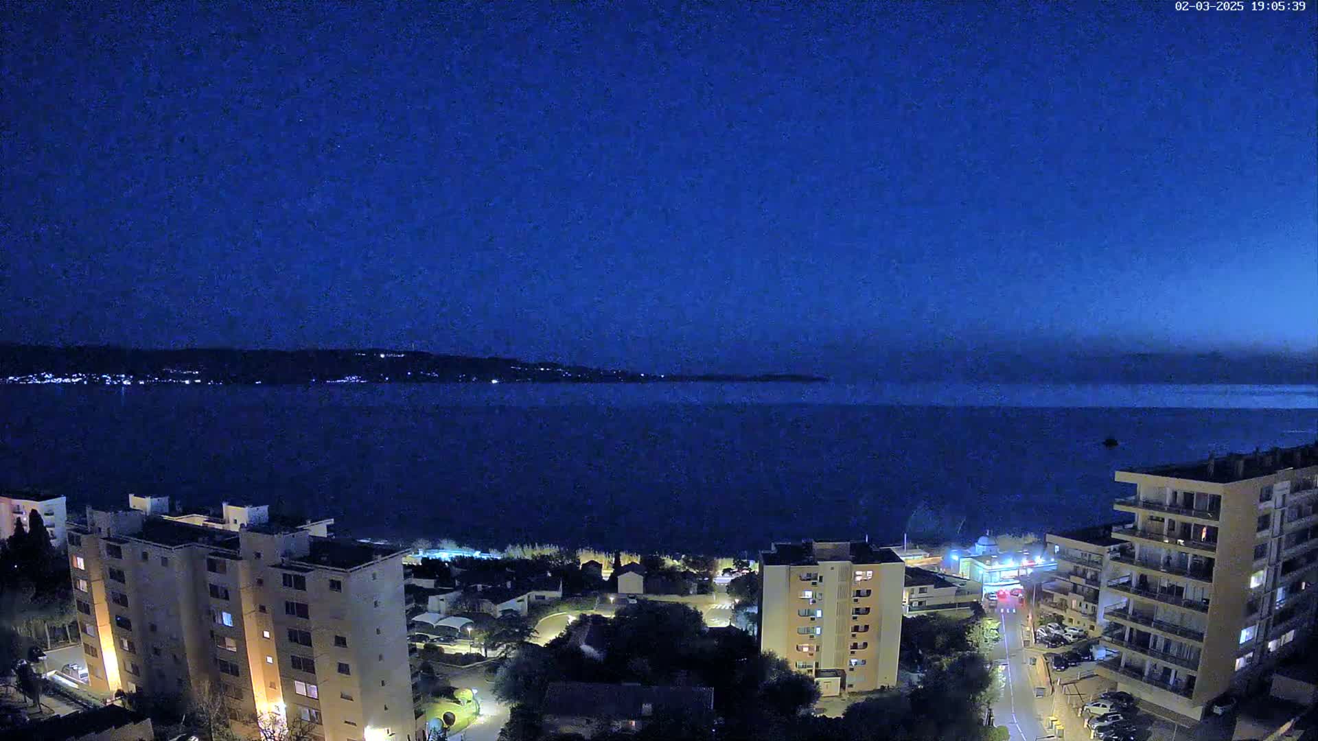 A nighttime view of a calm body of water, with a cityscape featuring several multi-story buildings along the shoreline under a dark blue sky.