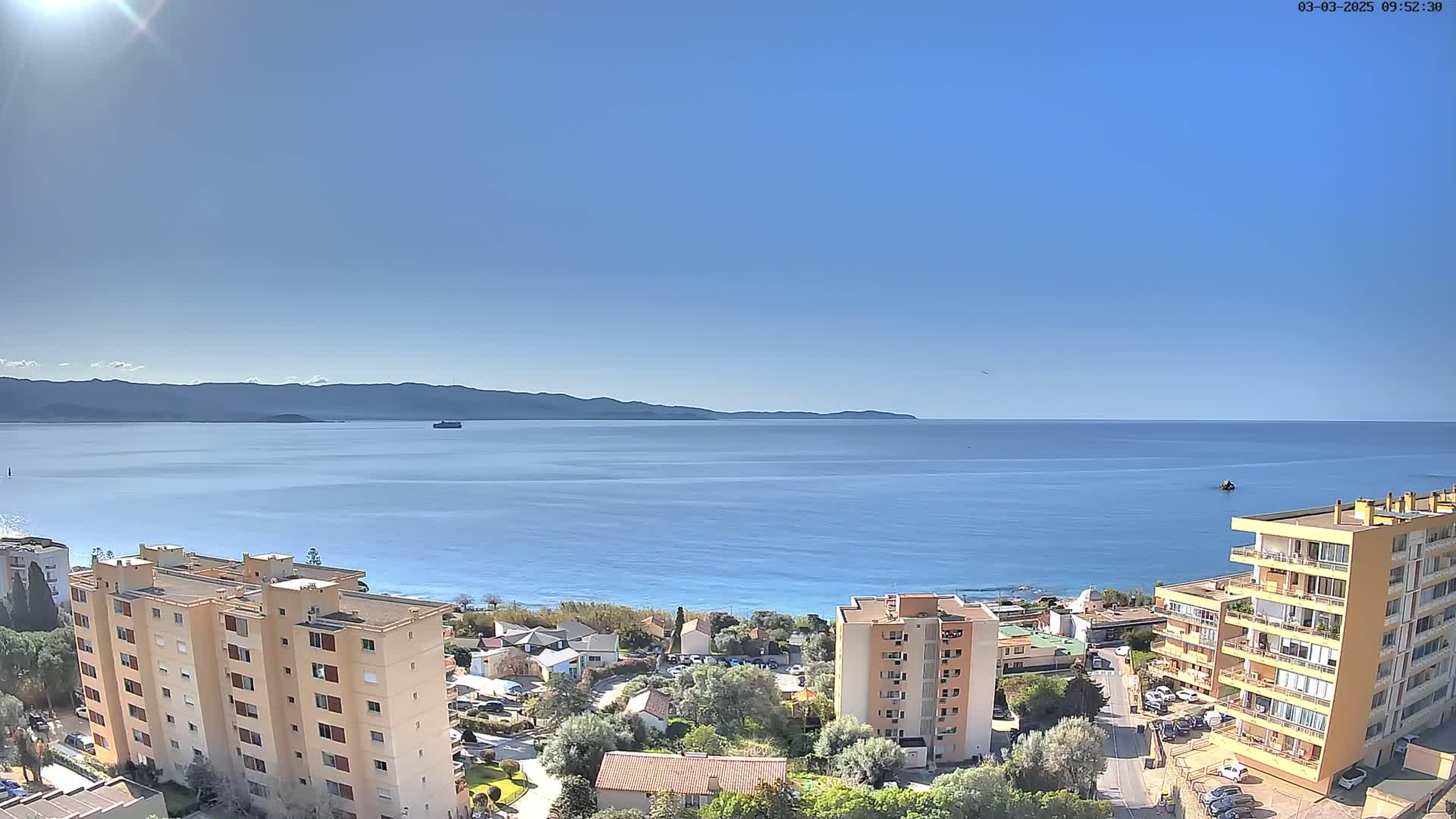 A sunny day reveals a coastal cityscape with apartment buildings overlooking a calm blue sea and distant mountains.