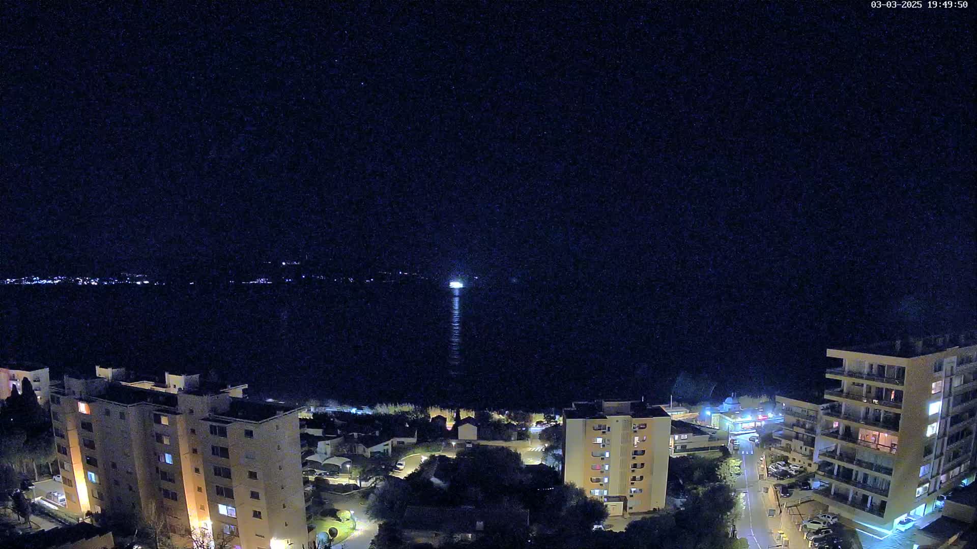 A nighttime, high-angle view shows a body of water with a light source in the distance, bordered by several illuminated apartment buildings and residential areas.