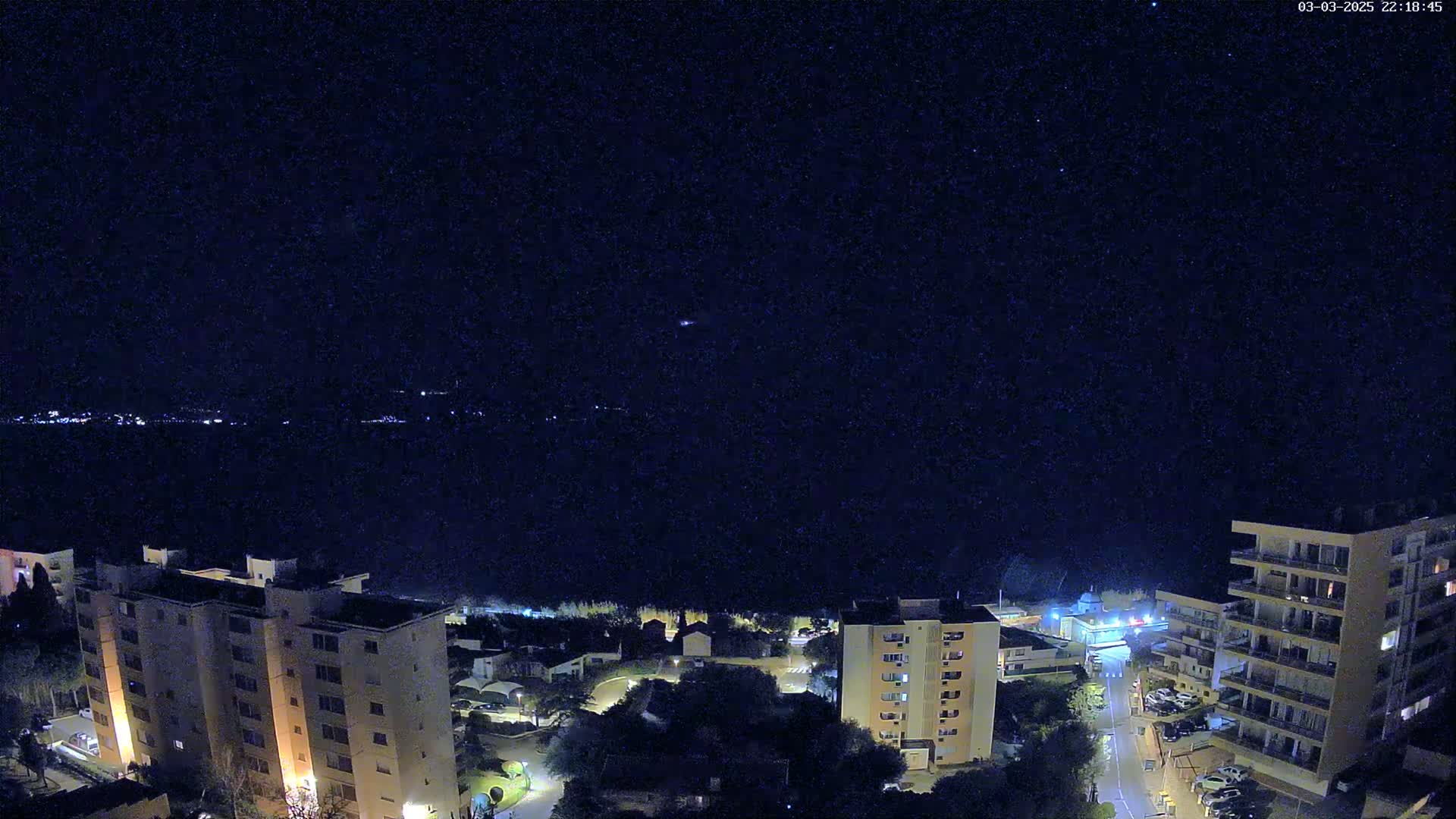 A nighttime aerial view shows several illuminated apartment buildings overlooking a dark body of water under a clear, starry sky.