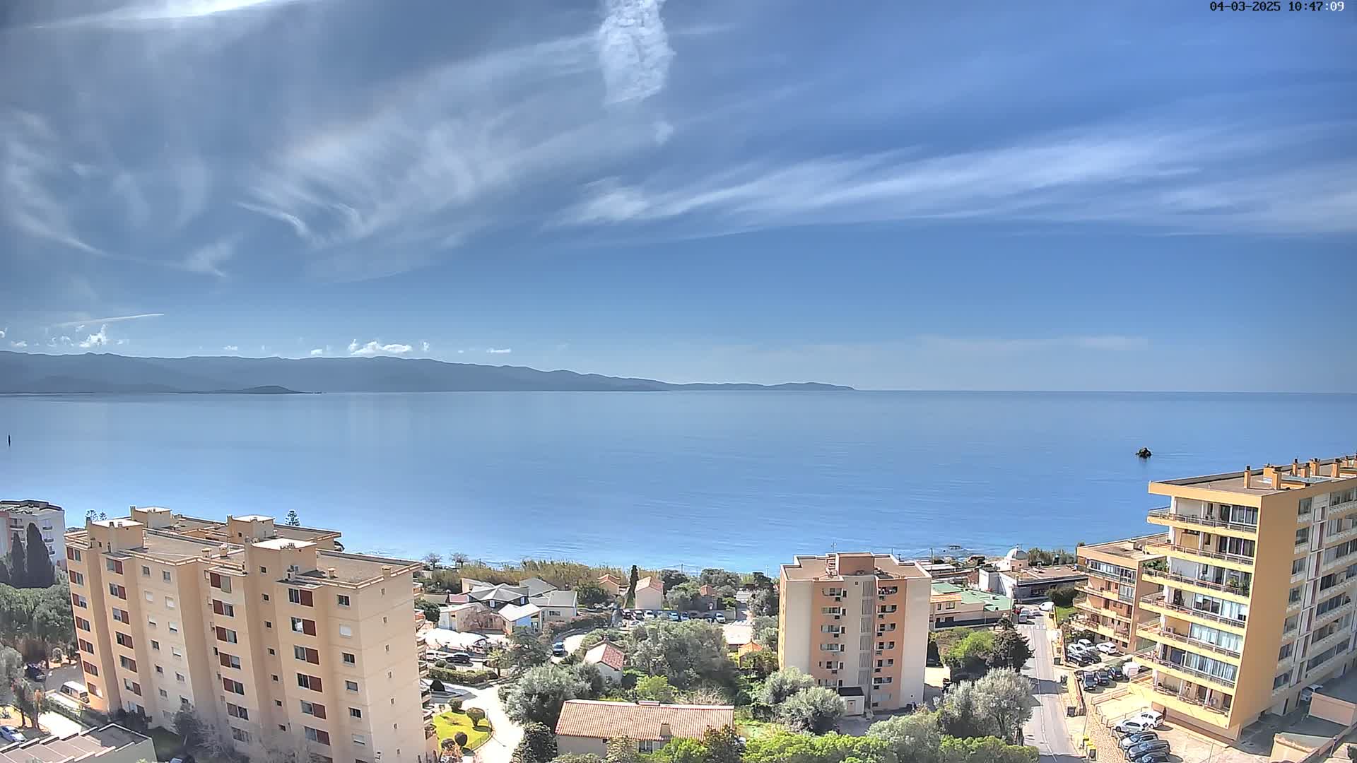 A mostly sunny day shows a calm blue sea stretching to distant mountains, with several apartment buildings and smaller homes along the shore.