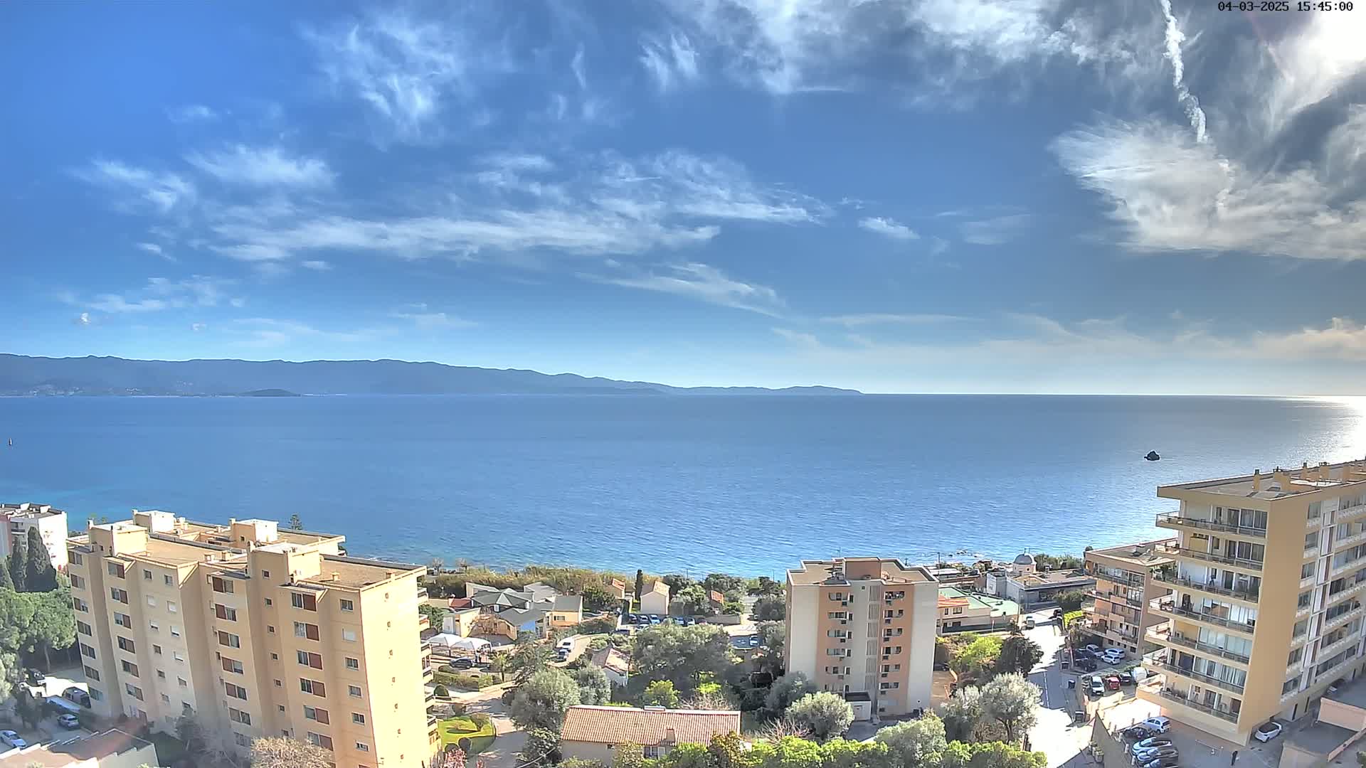 A mostly sunny day overlooks a calm blue sea with distant mountains under a partly cloudy sky, showing apartment buildings and roads nearby.