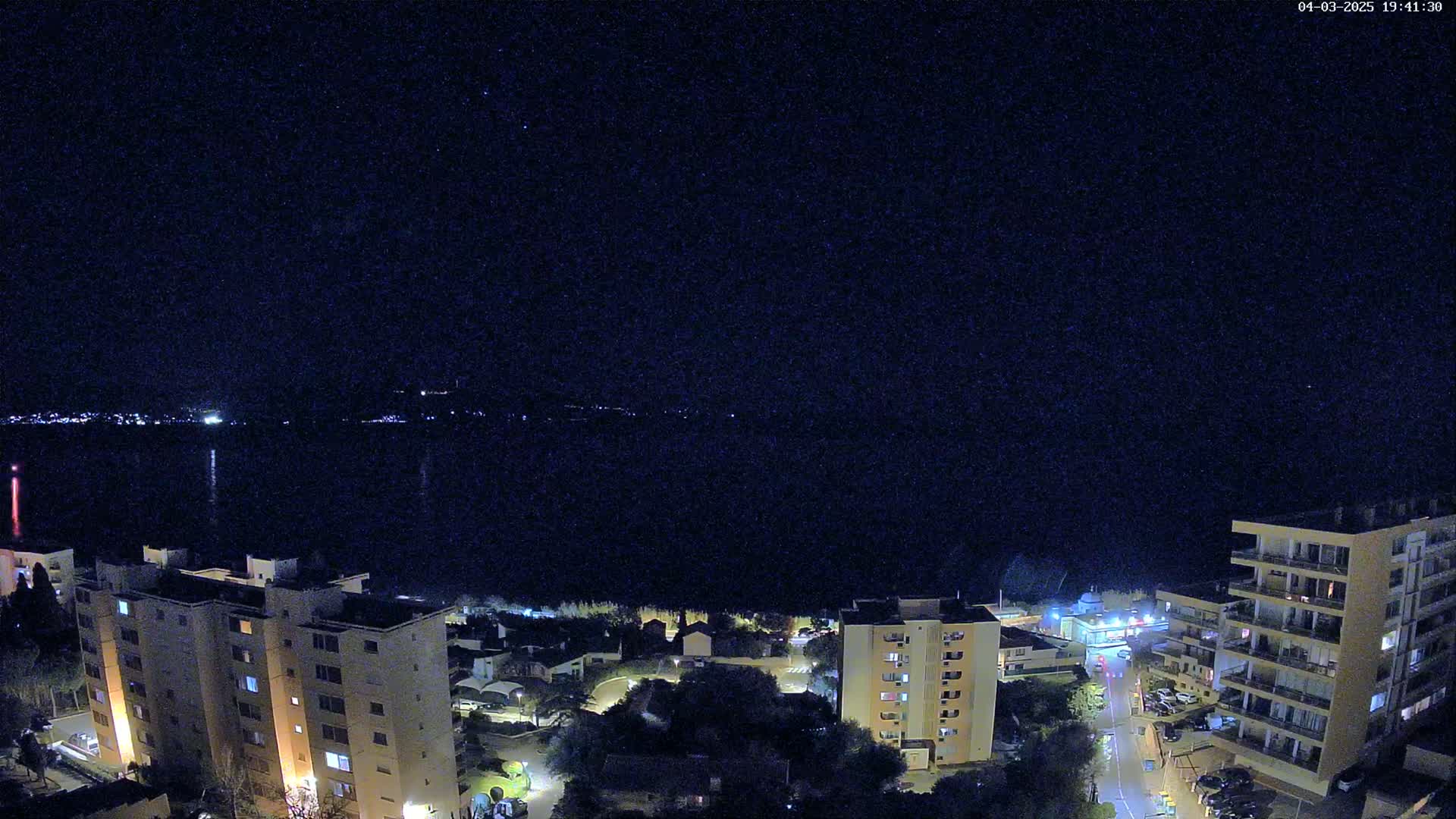 A nighttime aerial view shows a dark body of water with a city skyline and several apartment buildings illuminated by lights under a clear, starry sky.