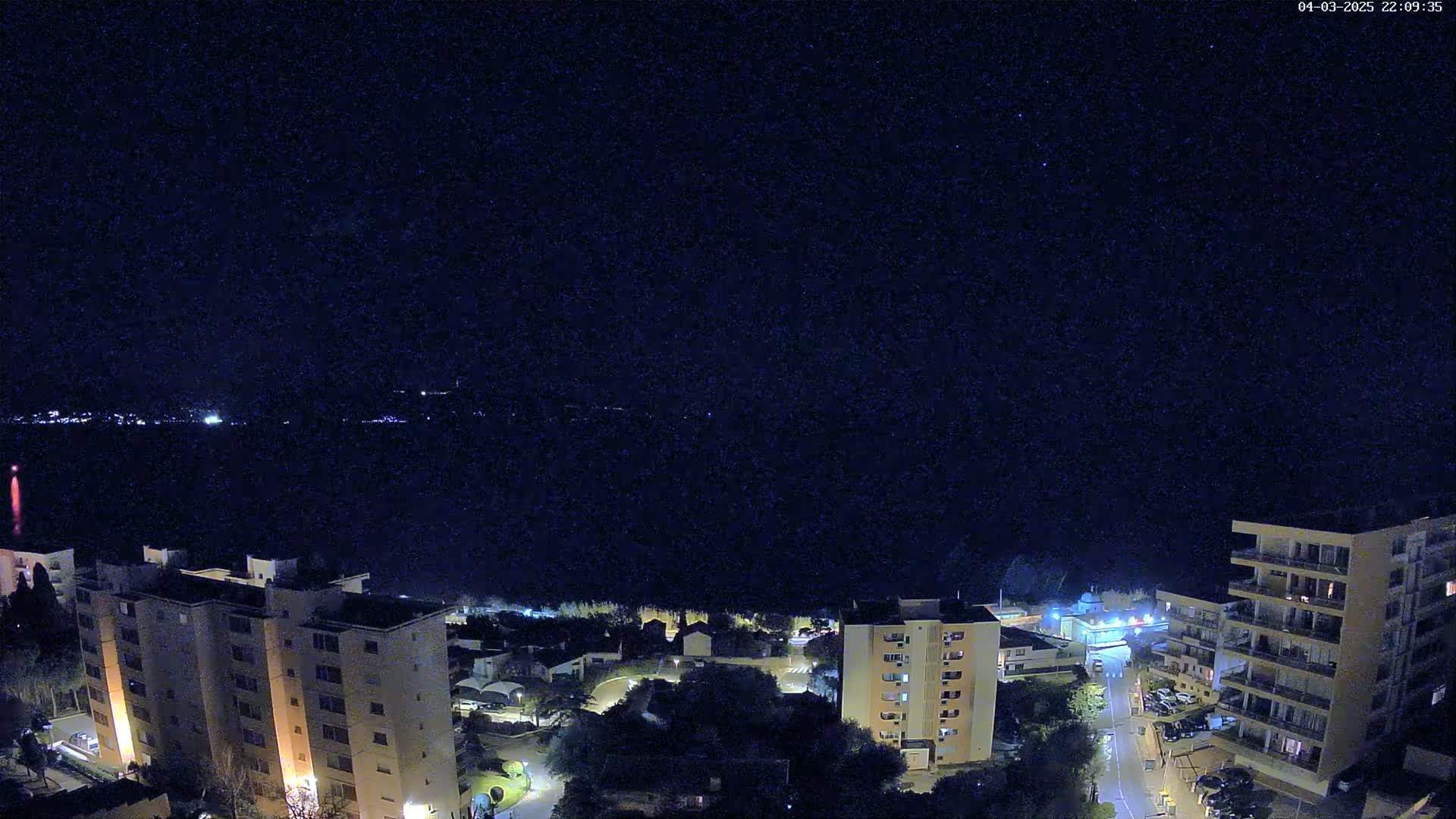 A night-time, high-angle view shows a body of water with city lights across it, and a collection of buildings with lit windows below.
