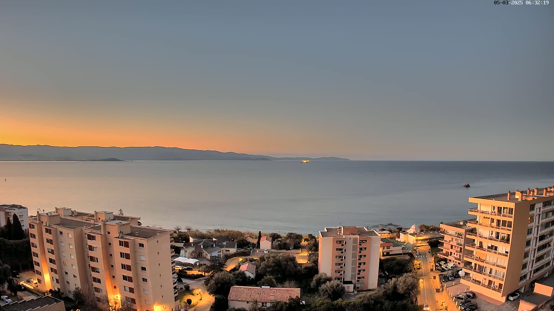 A coastal town at sunset, with calm water and several multi-story buildings illuminated, overlooks a tranquil sea and distant mountains.