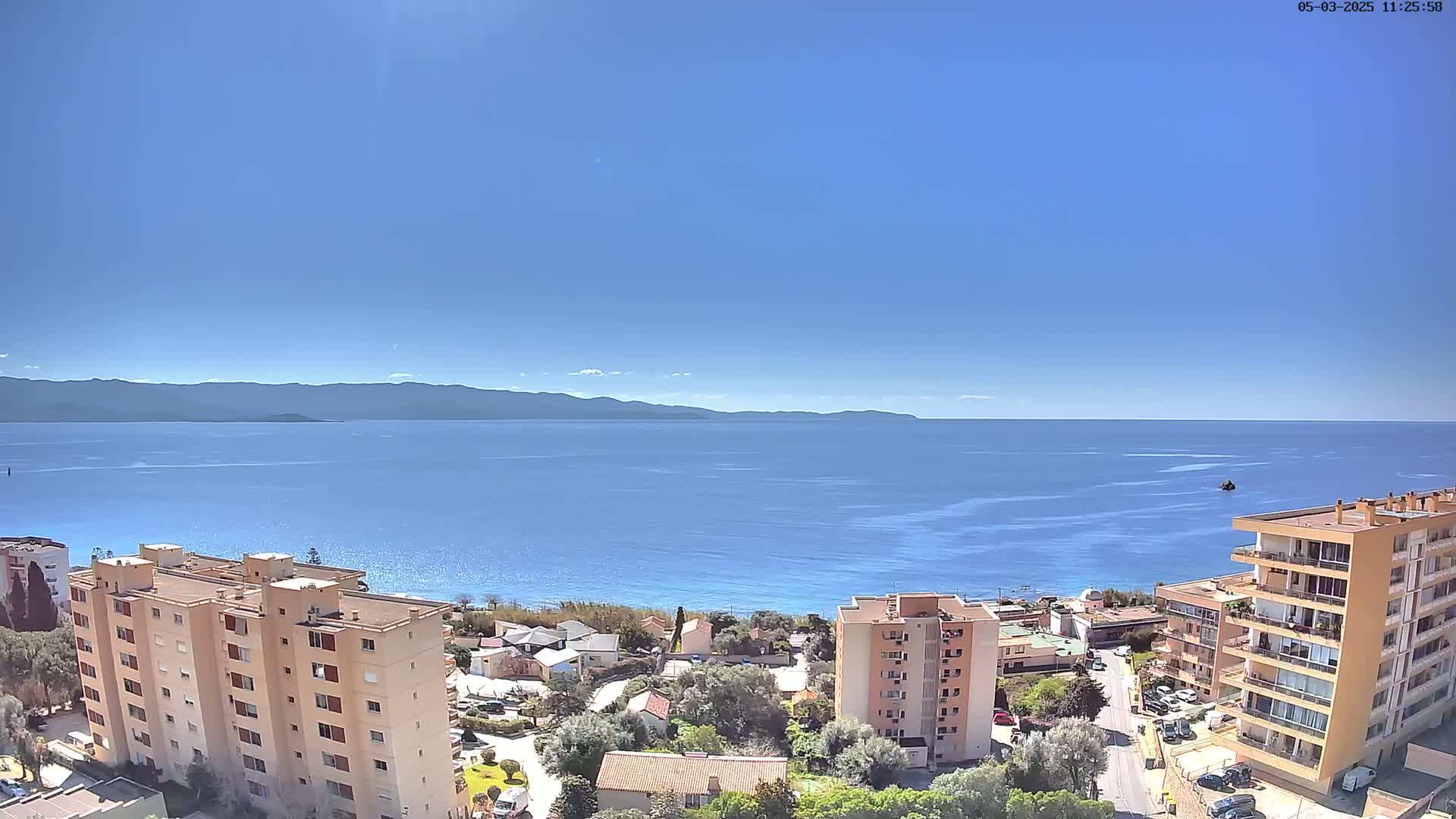 A sunny day reveals a coastal town with apartment buildings overlooking a calm blue sea and distant mountains under a clear blue sky.