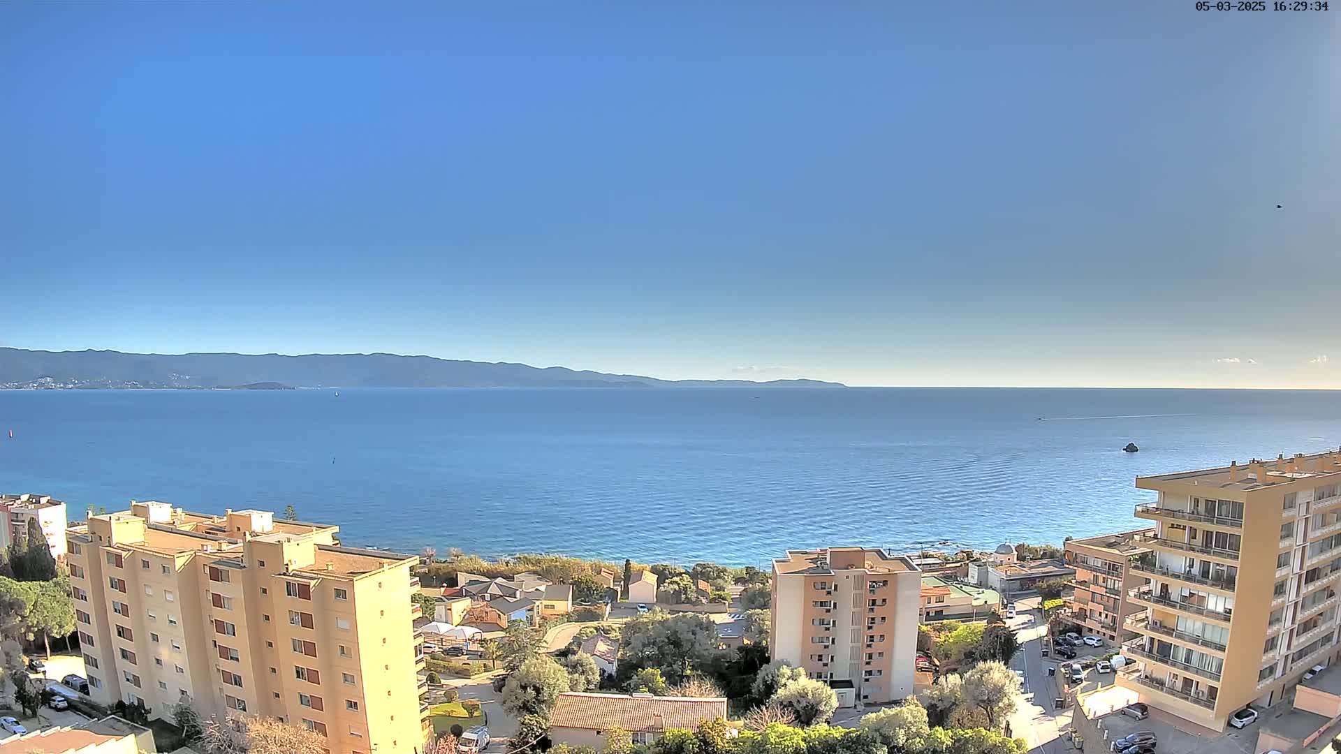 A clear, sunny day reveals a coastal town nestled between a calm blue sea and distant mountains.