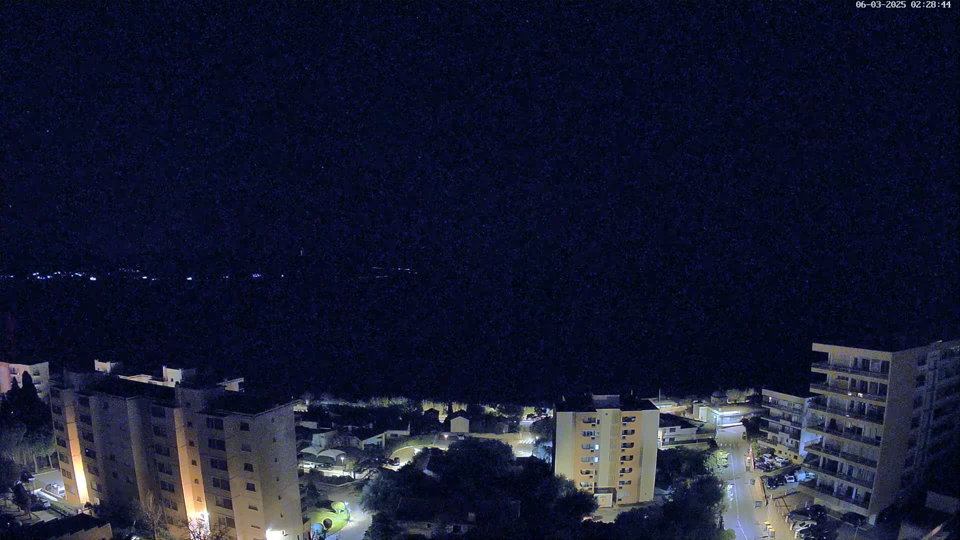 A nighttime aerial view shows several apartment buildings illuminated, nestled among darker trees and roads under a clear, dark sky.