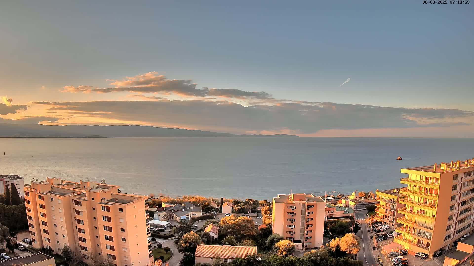 An aerial view shows a calm sea at sunset, with several apartment buildings and other structures along a coastal hillside.