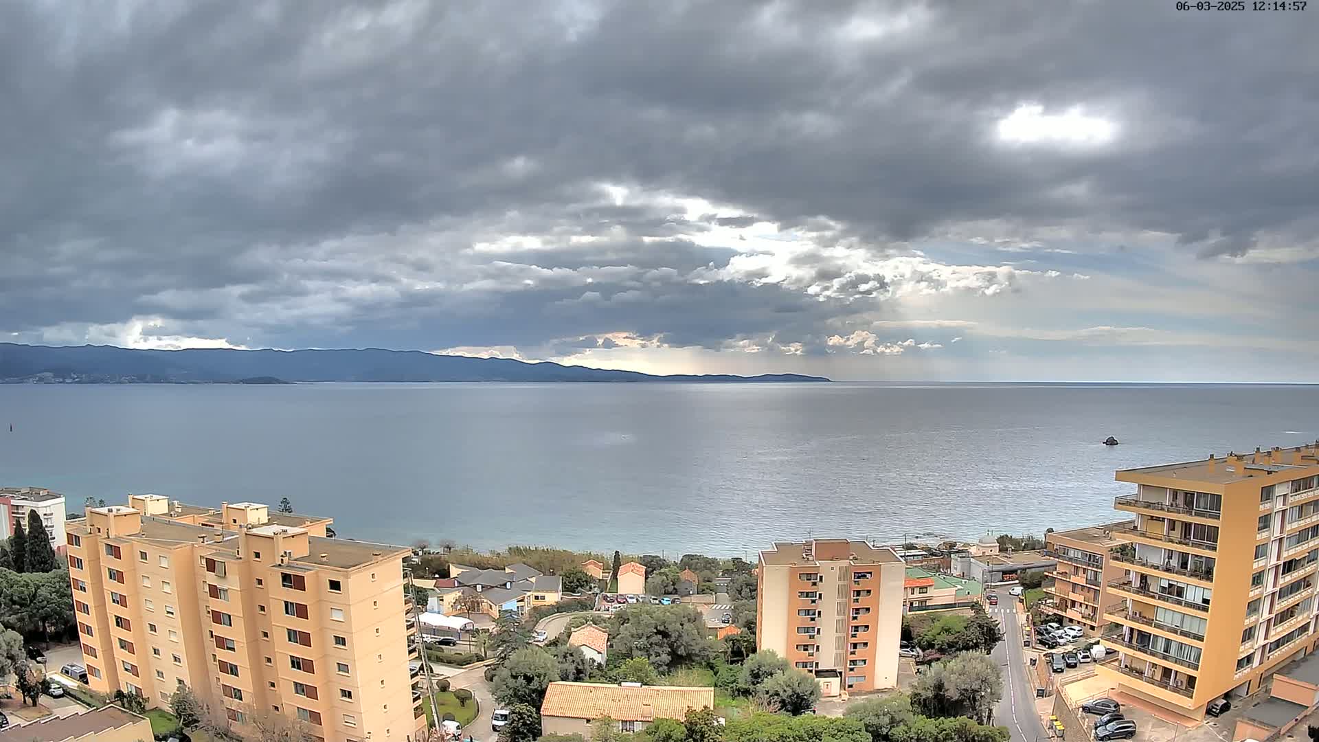 An overcast sky hangs over a calm sea, with a view of coastal buildings and distant mountains.