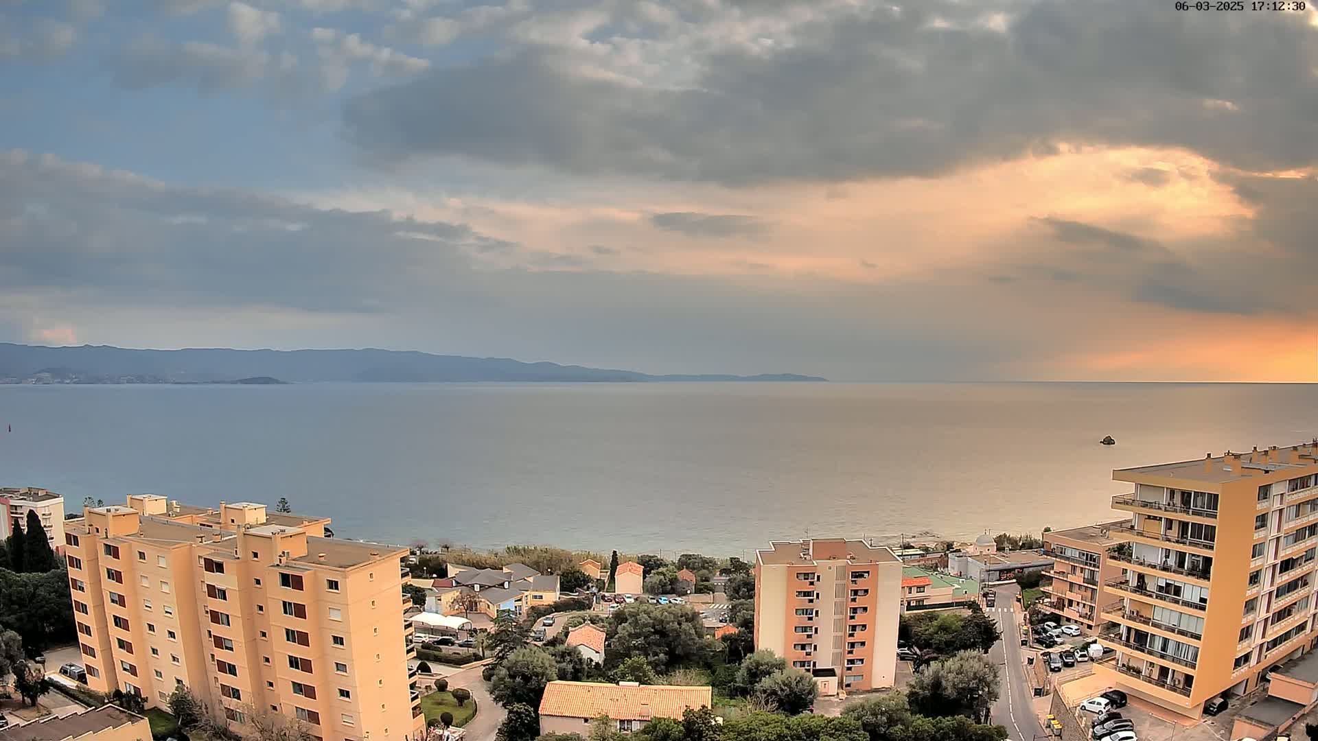 An elevated view shows a calm sea under a partly cloudy sunset sky, with several apartment buildings and a coastal town nestled between the water and distant mountains.