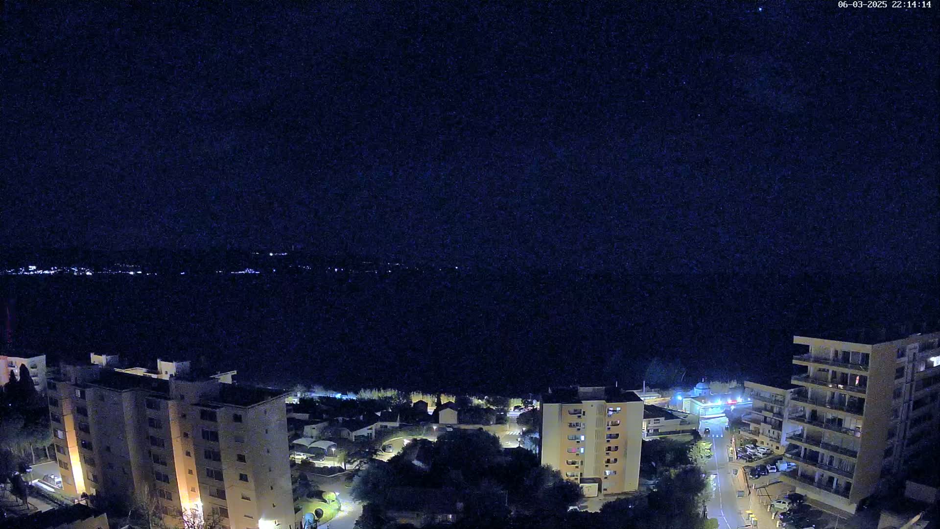 A nighttime aerial view shows a city overlooking a dark body of water under a clear, starry sky.
