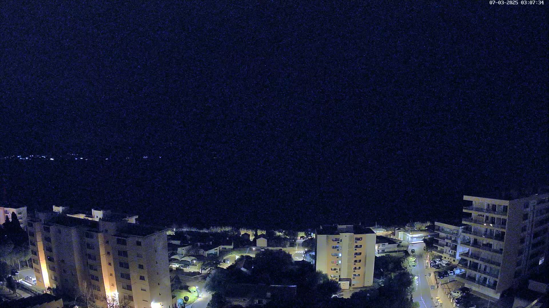 A nighttime aerial view shows several apartment buildings and residential houses illuminated, with a dark body of water in the distance under a clear, dark sky.