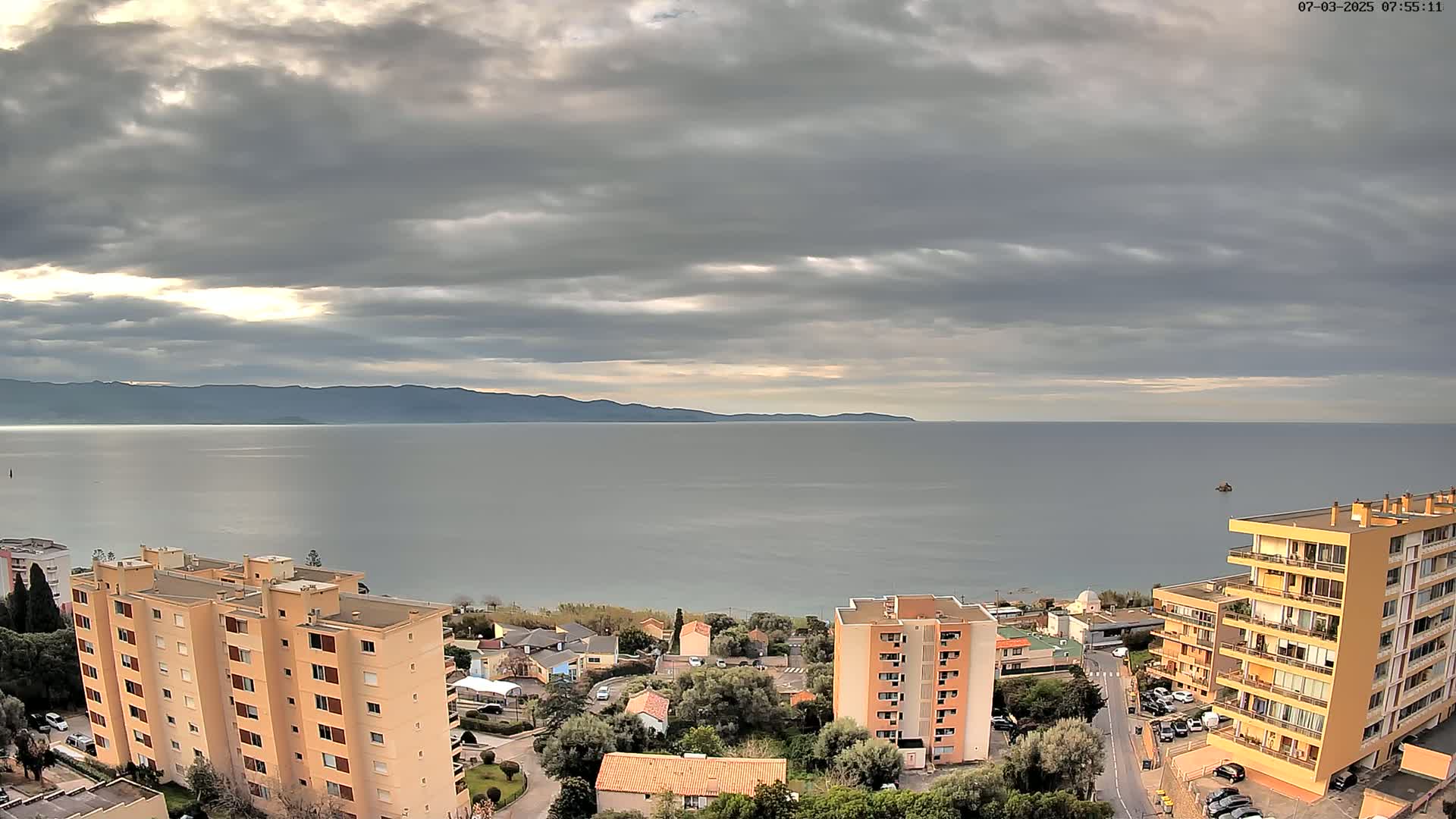 A coastal town with apartment buildings is viewed from above under a cloudy sky, overlooking a calm sea and distant mountains.