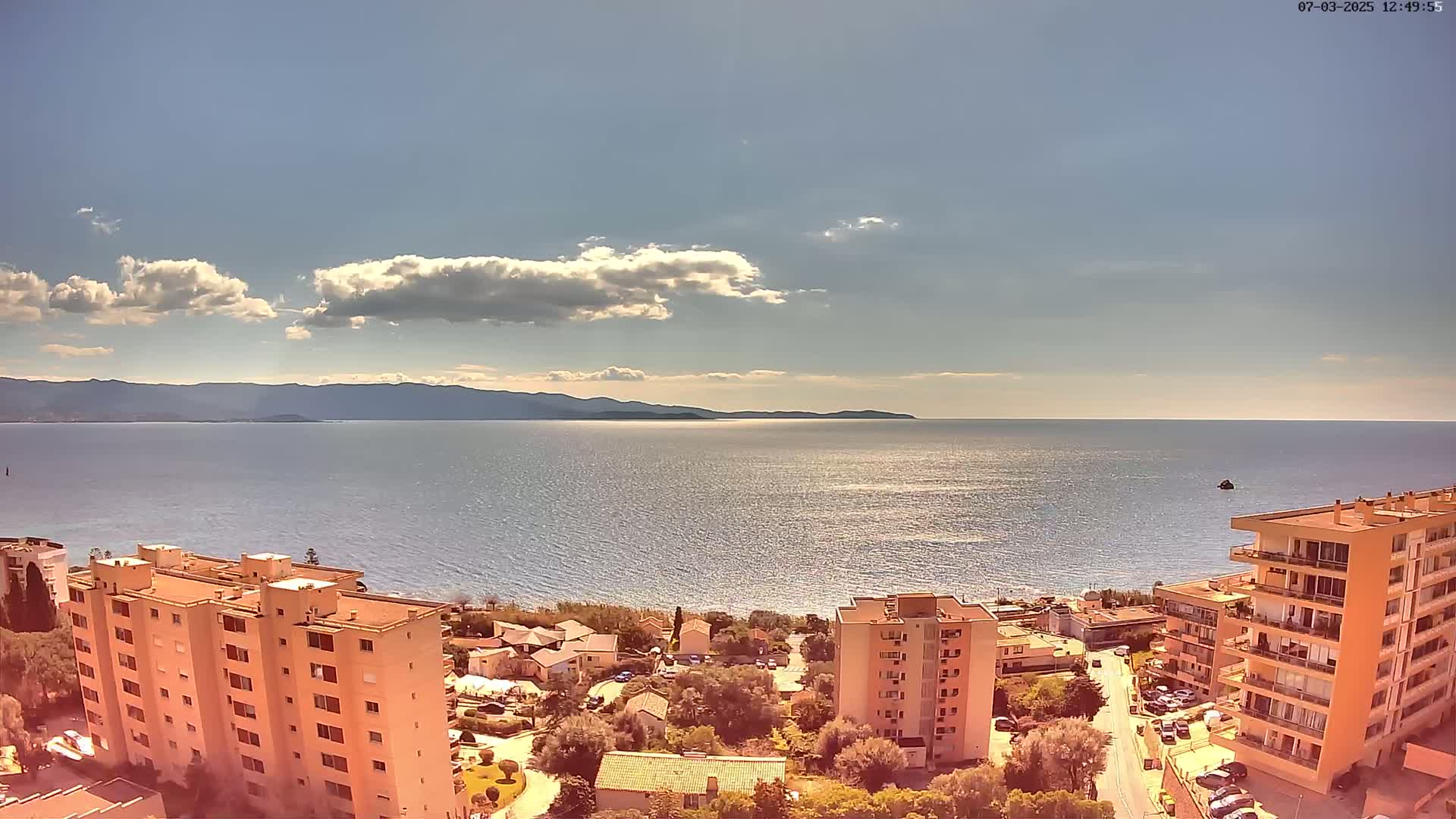 An aerial view shows a partly sunny day overlooking a calm sea with distant mountains, and several multi-story buildings situated near a shoreline with low-lying houses.