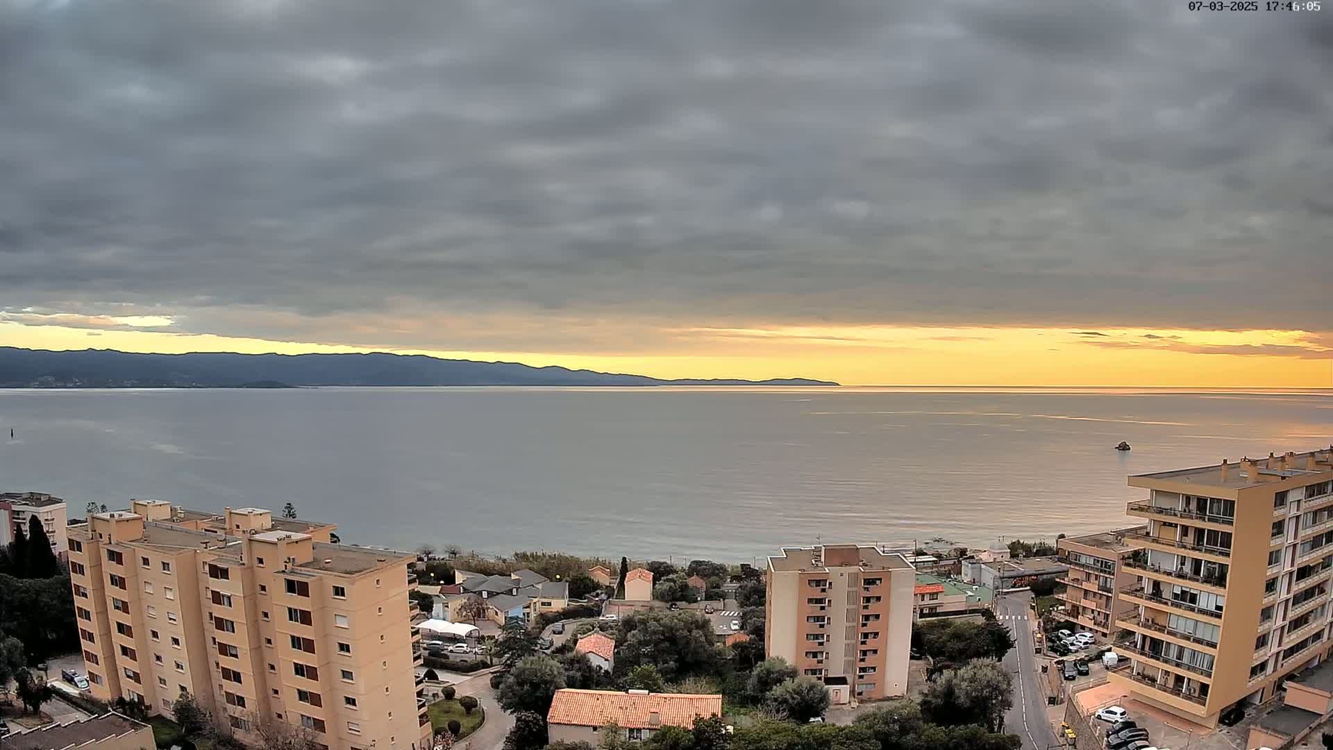 A coastal sunset view shows a calm sea under a partly cloudy sky, with several apartment buildings overlooking the water and a mountain range in the distance.