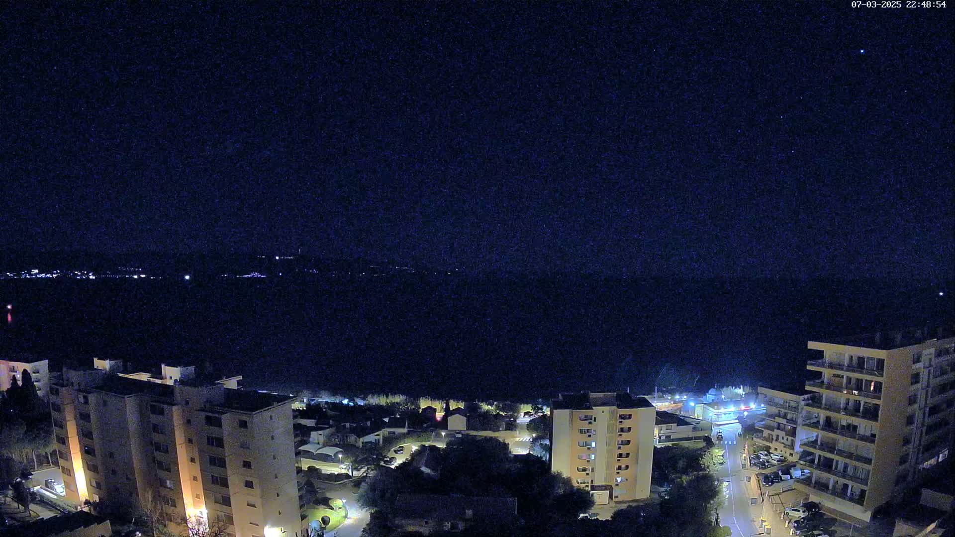 A nighttime view of a coastal town with apartment buildings and a starry sky over calm water.