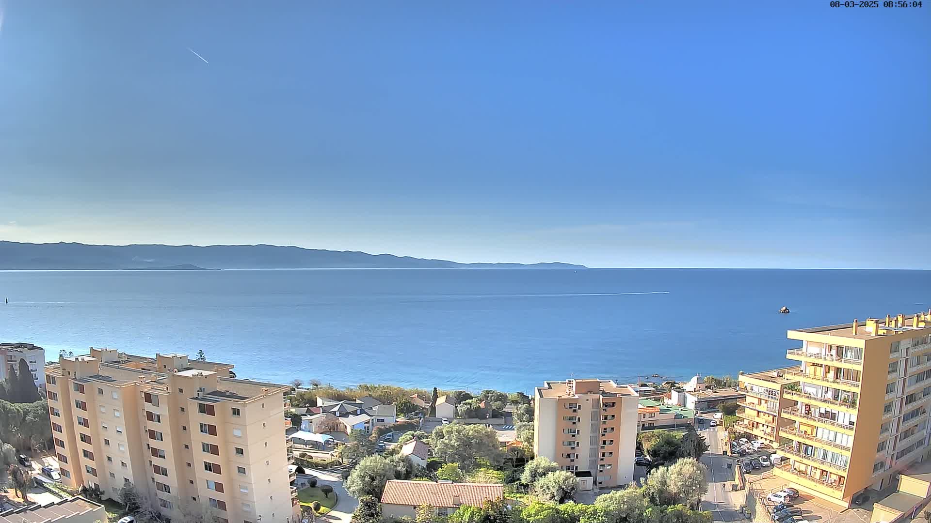 A clear, sunny day overlooks a calm blue sea with distant mountains, and several multi-story buildings situated along the coast.