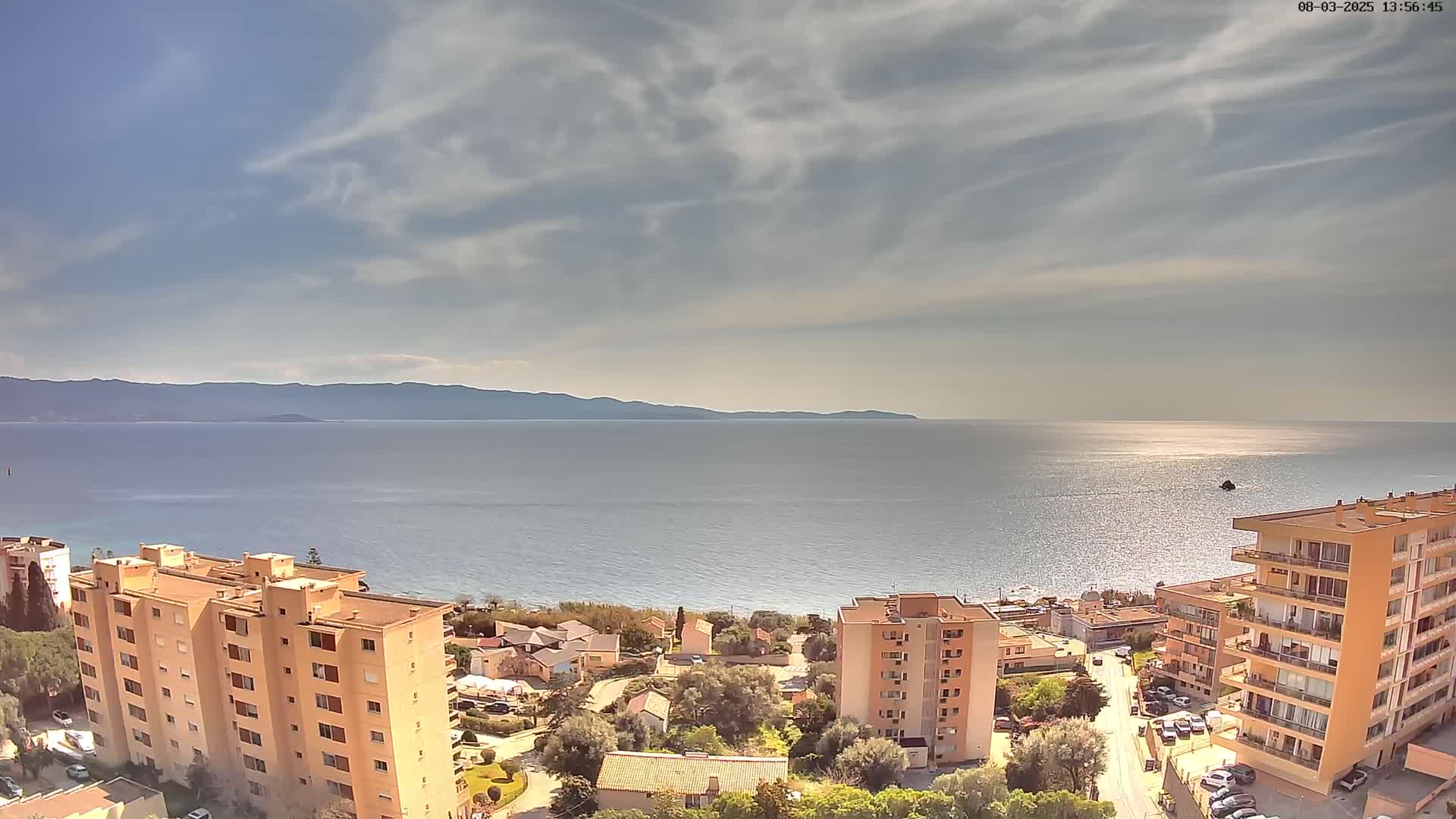 A partly cloudy sky overlooks a calm sea with a distant mountain range, and several multi-story buildings and houses situated along a shoreline.
