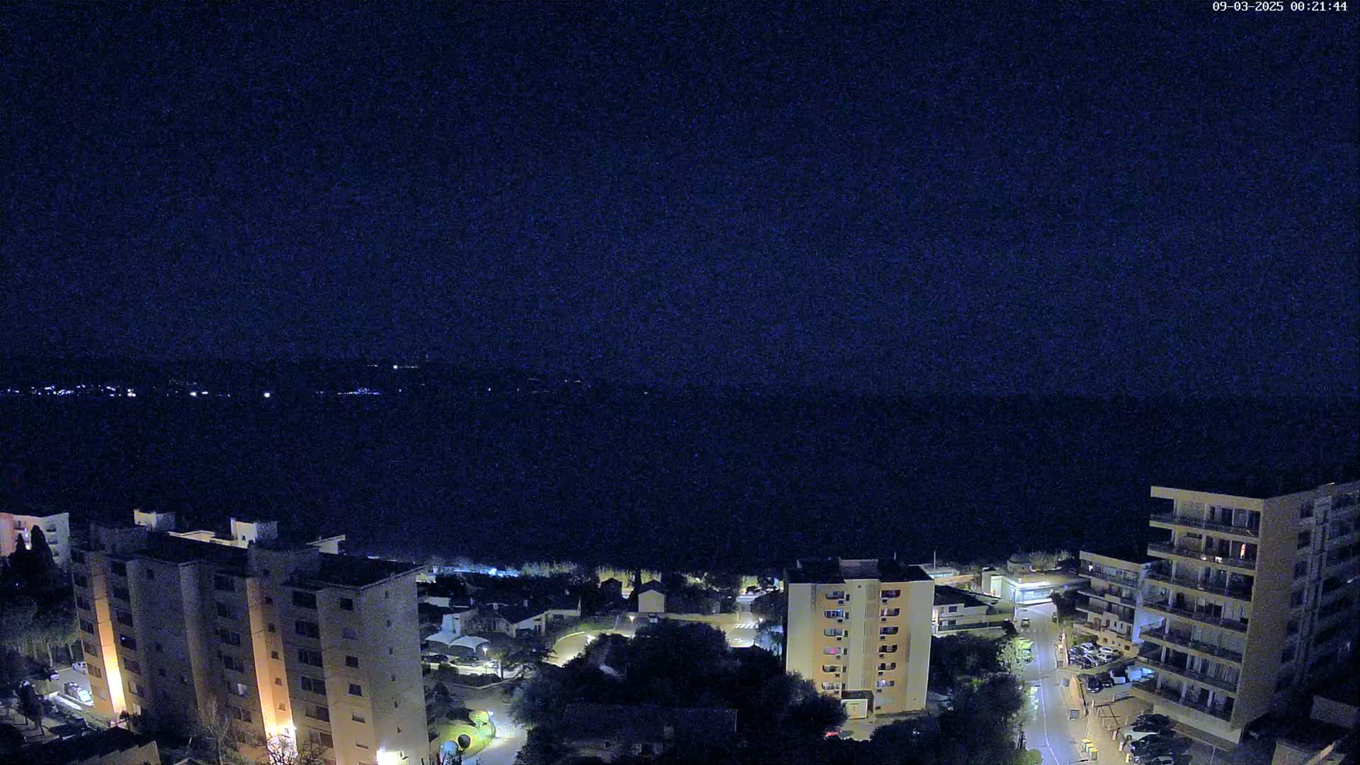 A nighttime aerial view shows several illuminated apartment buildings overlooking a dark body of water under a clear, dark sky.