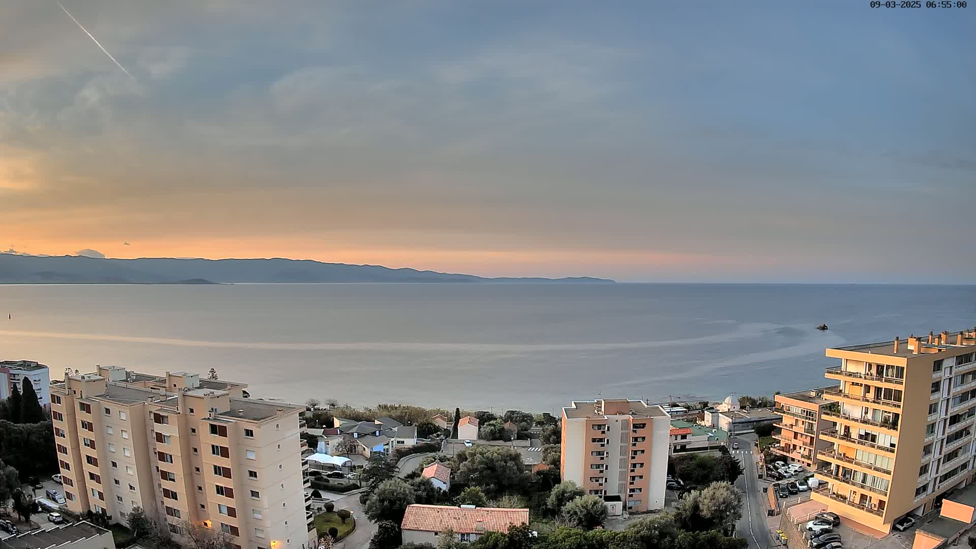 An elevated view shows a calm sea under a mostly cloudy, pastel-colored sky at sunset, with several apartment buildings and a coastal town nestled below, backed by distant mountains.