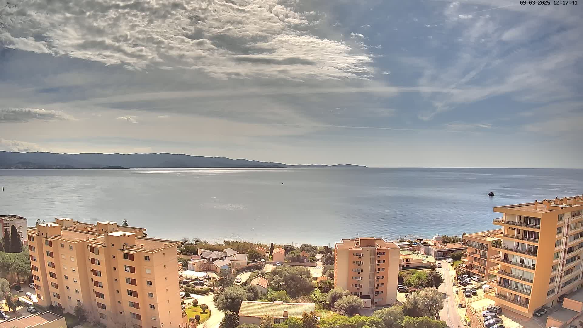 A partly cloudy day reveals a calm sea extending to distant mountains, with several apartment buildings and houses situated along a shoreline.