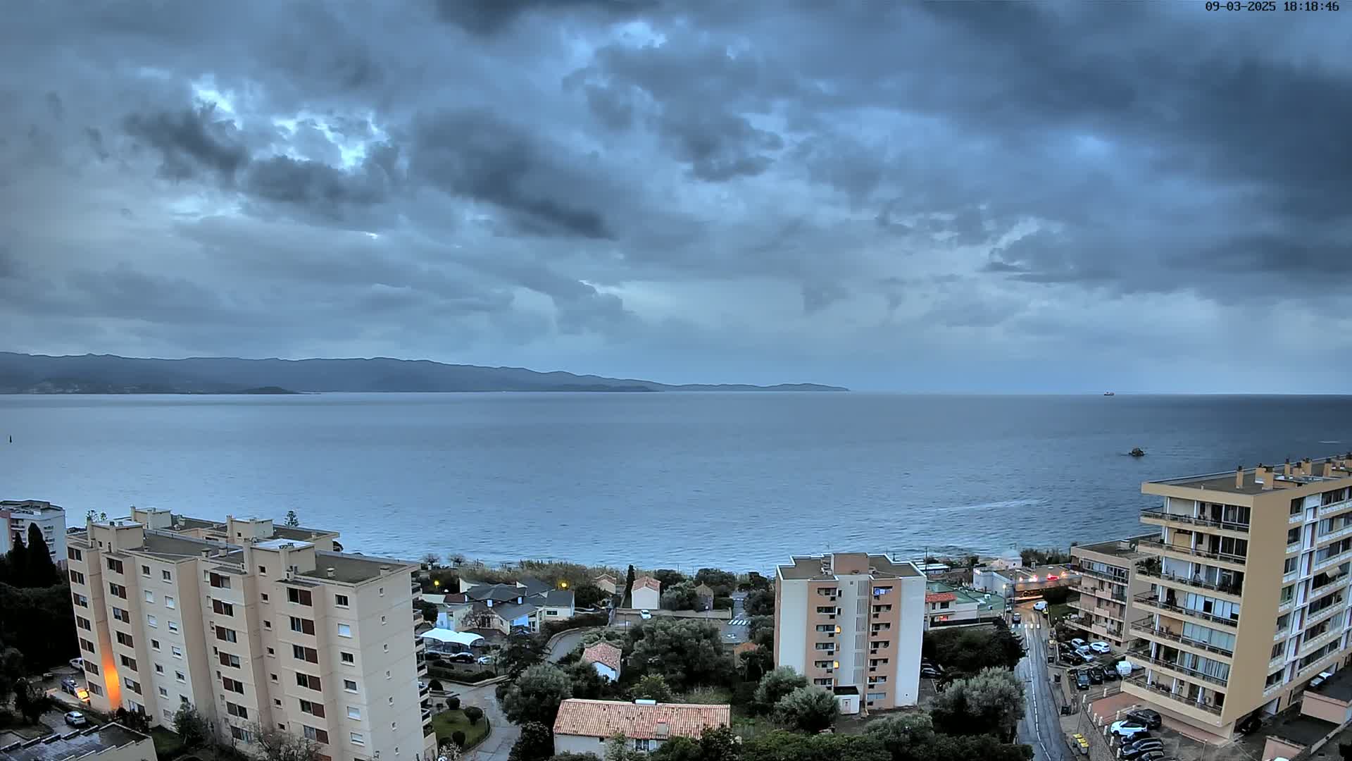 An overcast sky hangs over a calm sea, overlooking coastal buildings and a distant mountain range.