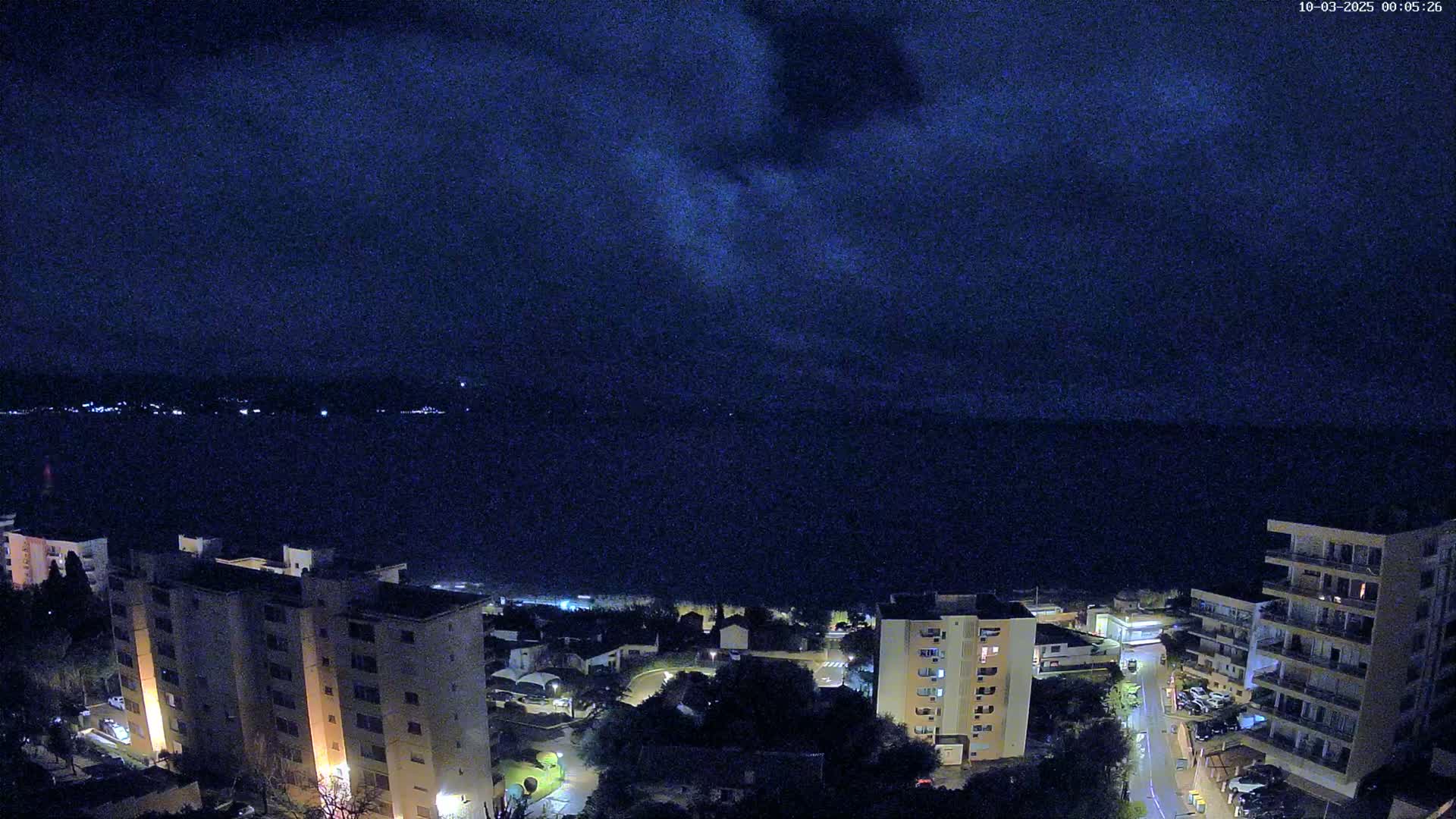 A nighttime view of a coastal town with apartment buildings illuminated, overlooking a dark body of water under a mostly clear, dark sky.