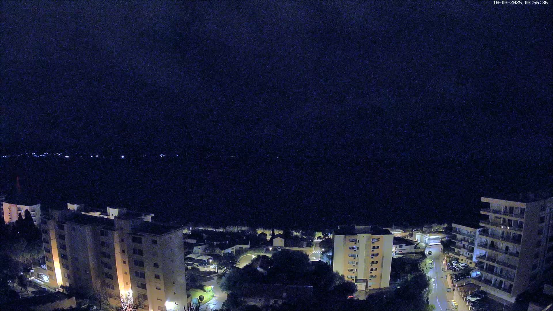 A nighttime aerial view shows a coastal town with several apartment buildings and a dark, clear sky.