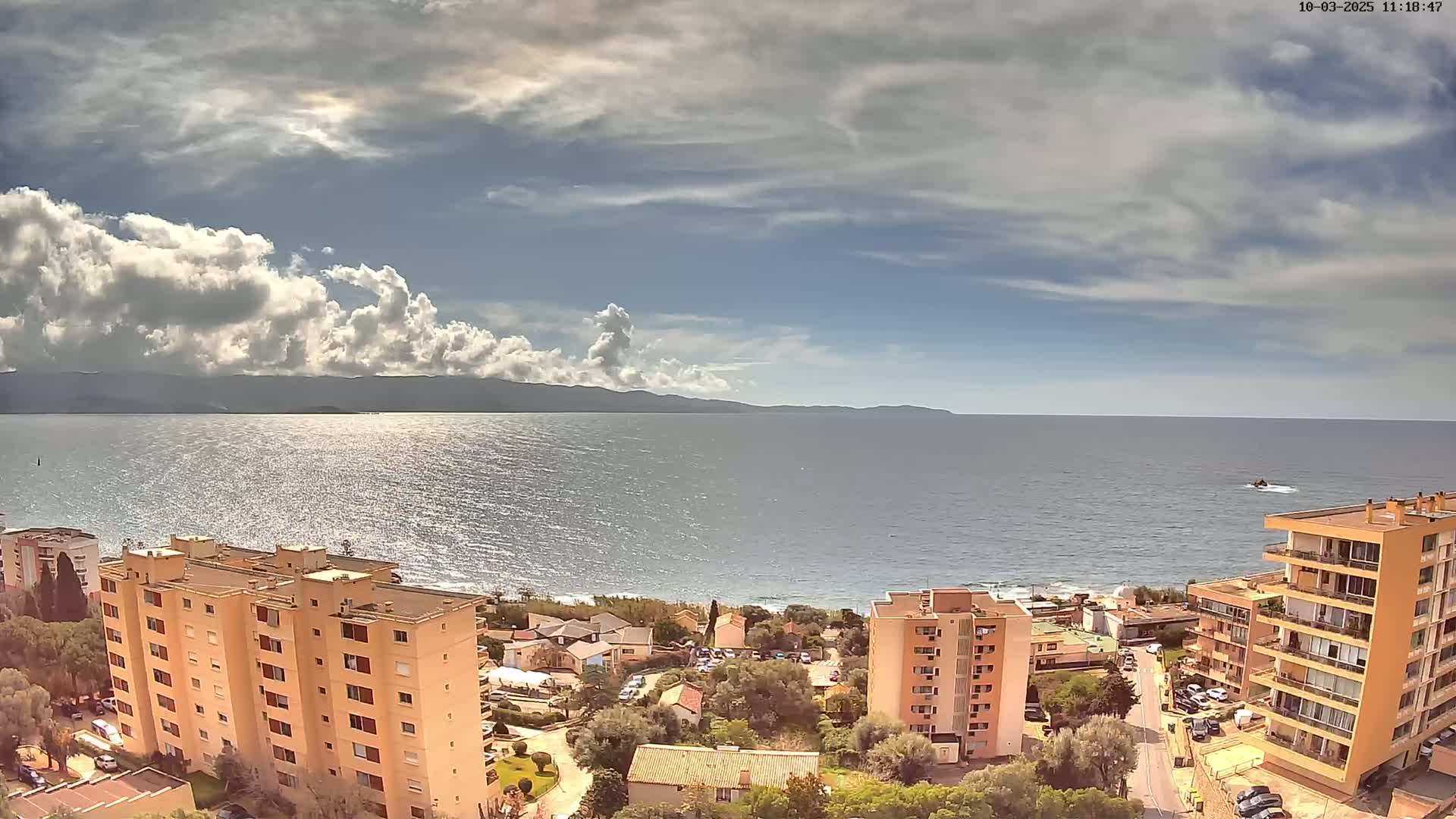 A partly cloudy day shows a view of a calm sea with buildings and vegetation near a shoreline, mountains in the distance.