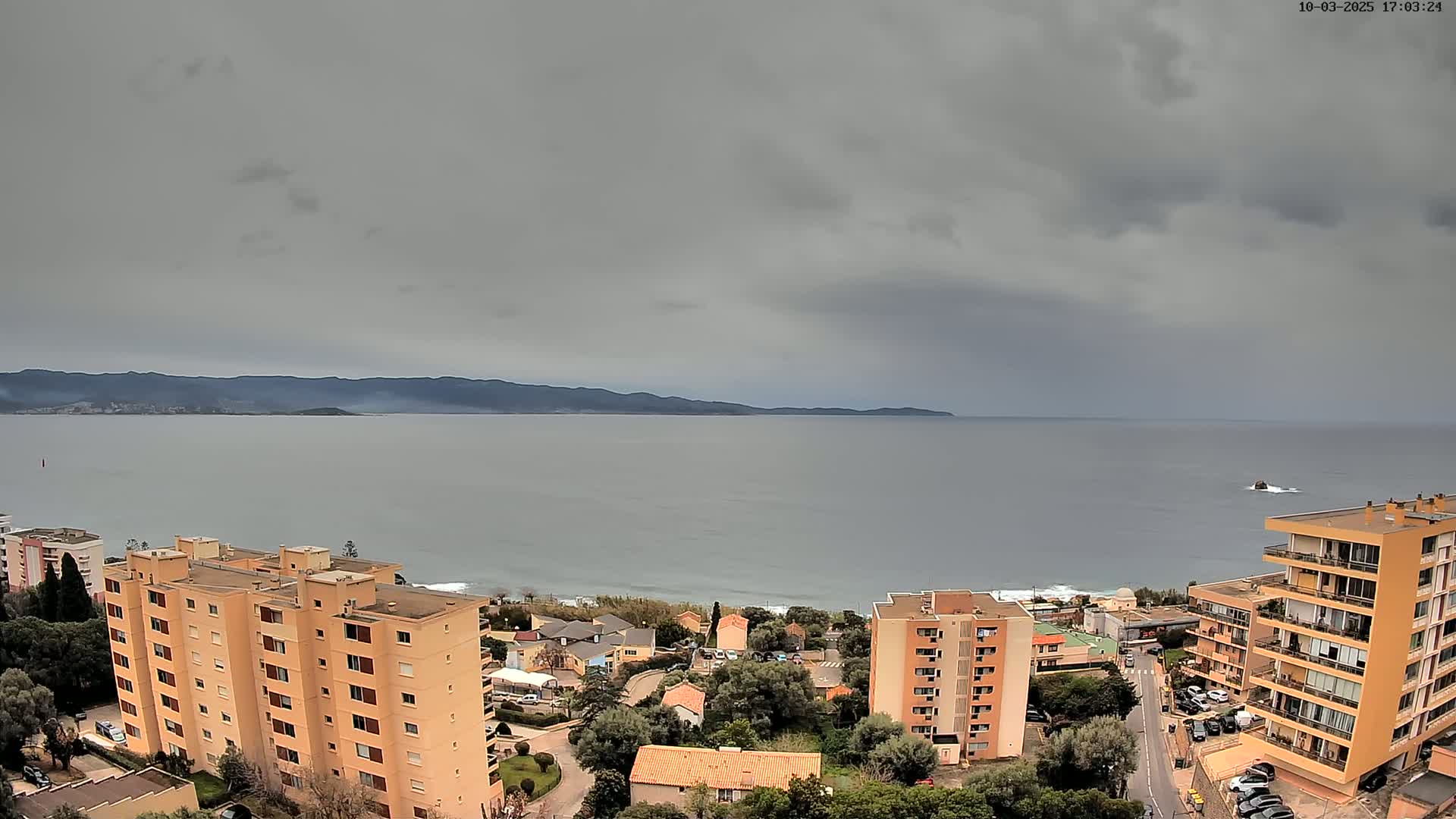 An overcast sky hangs over a calm sea, where a shoreline town with several apartment buildings sits nestled between the water and distant mountains.