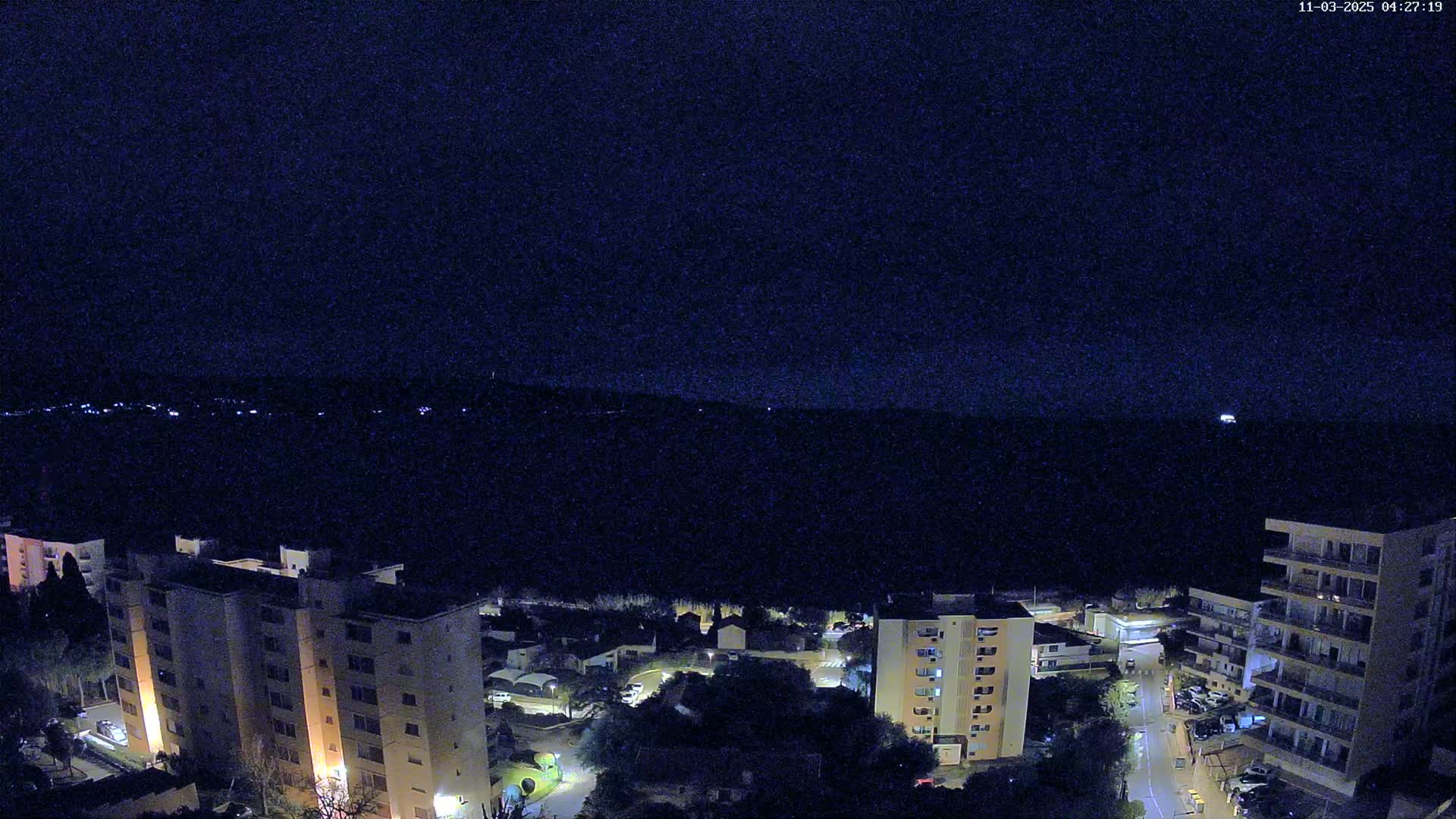 A nighttime, high-angle view shows several illuminated apartment buildings and a street, with a dark, distant horizon under a clear, dark sky.