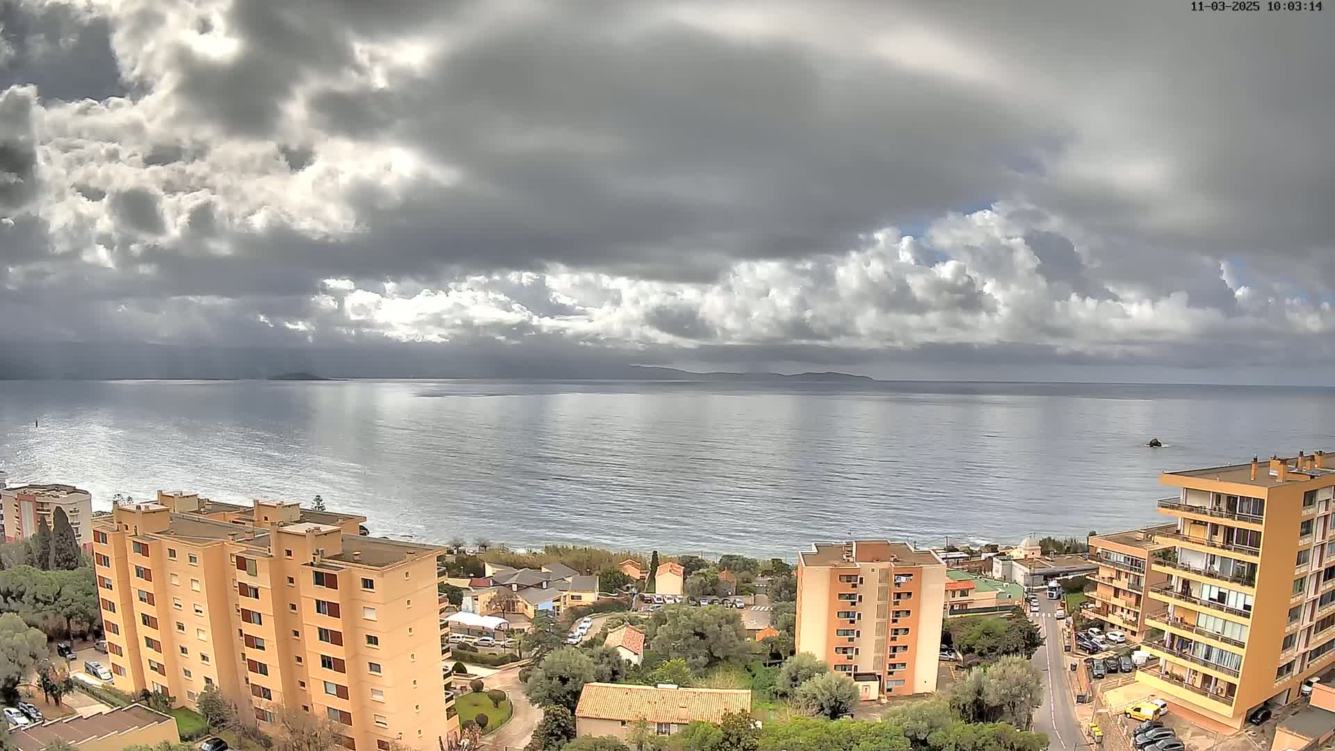 A partly cloudy day overlooks a calm sea, with several multi-story apartment buildings situated on a hillside overlooking the water.
