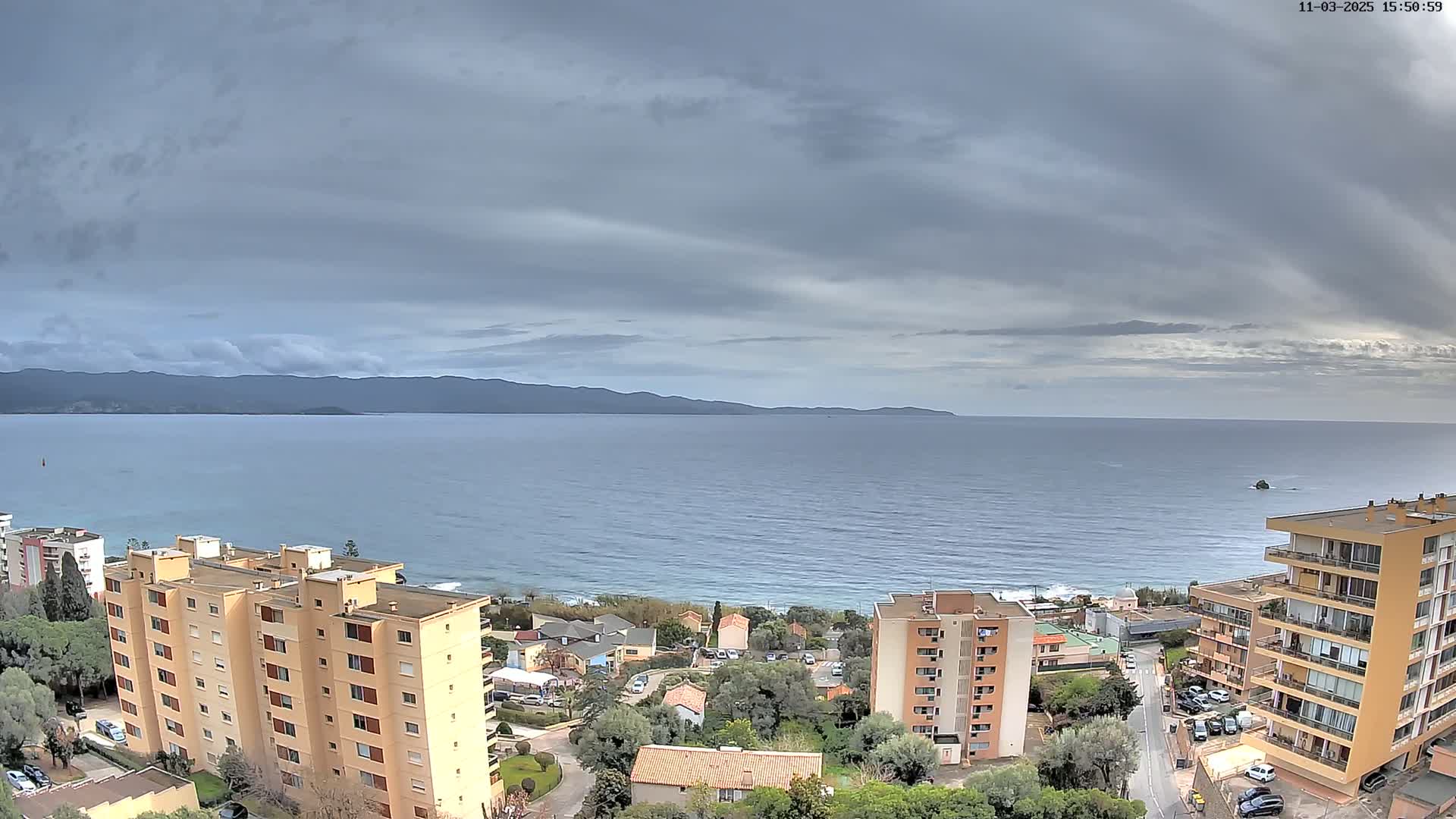 A coastal view under a cloudy sky shows a calm sea, distant mountains, and several apartment buildings nestled among trees and houses.