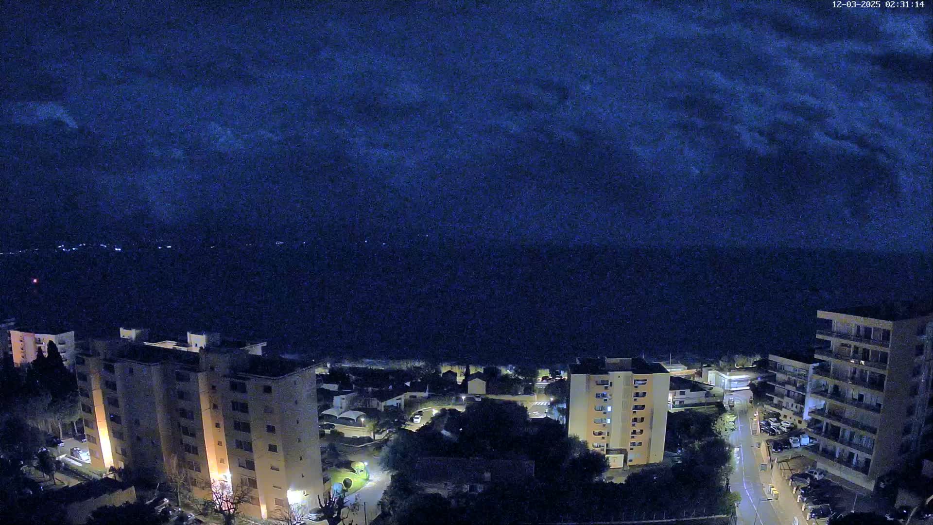 A nighttime view of a coastal town with apartment buildings and a dark, cloudy sky over a calm sea.