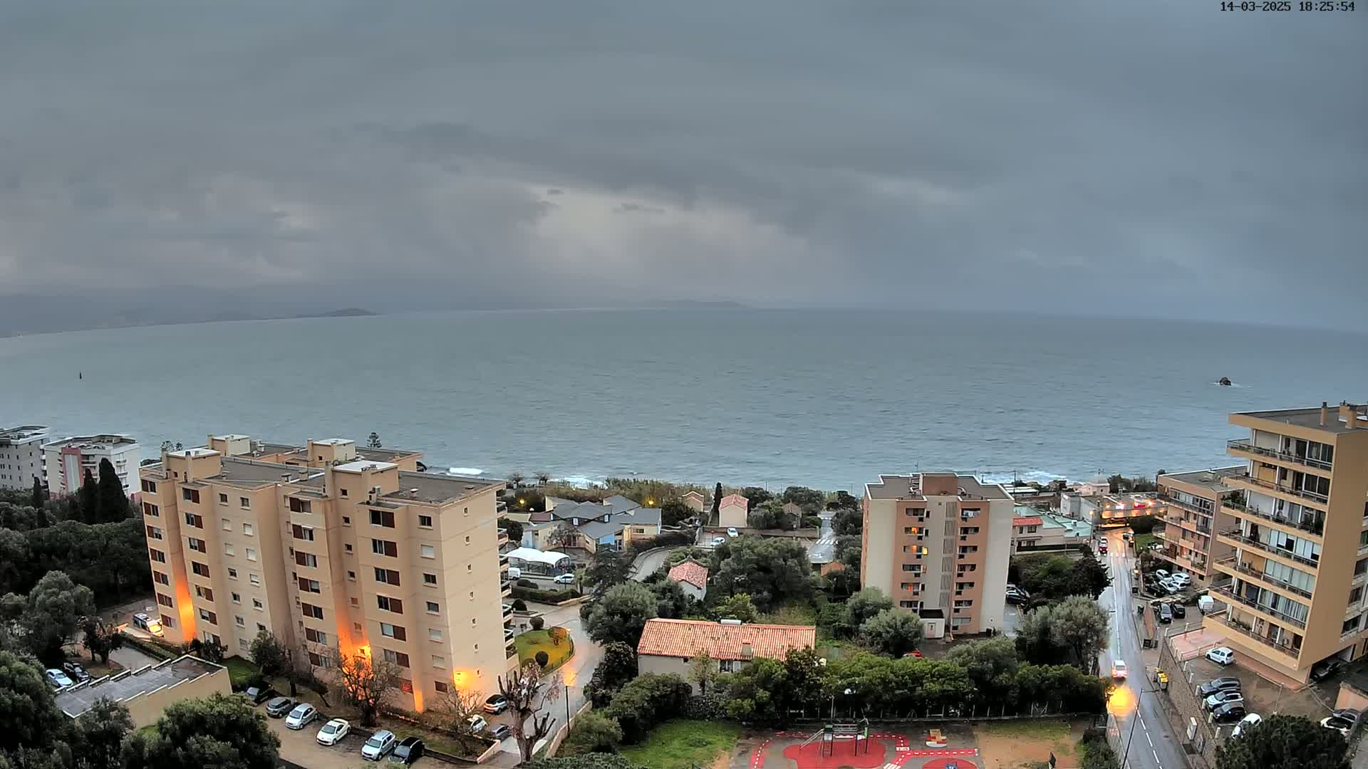 An overcast coastal town with several apartment buildings is seen from an elevated vantage point, overlooking a calm sea under a gray sky.