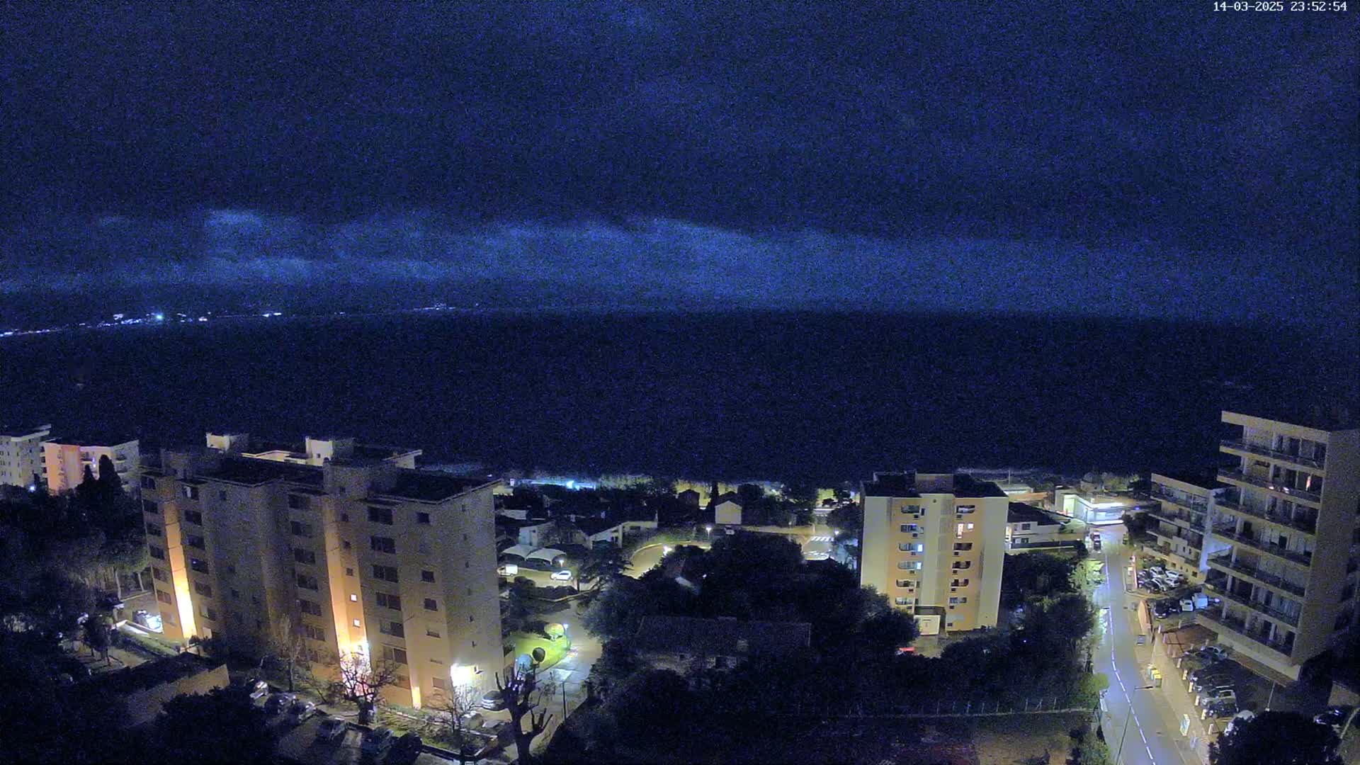 A nighttime view of a coastal town shows several apartment buildings illuminated against a dark, cloud-covered sky and a calm sea.