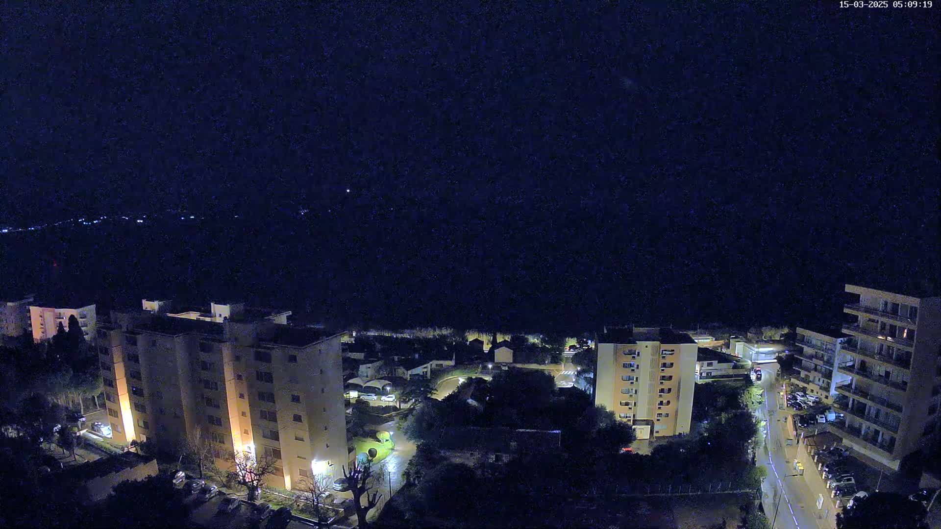 A nighttime, high-angle view shows several multi-story apartment buildings illuminated, surrounded by dark trees and a street with parked cars under a clear, dark sky.