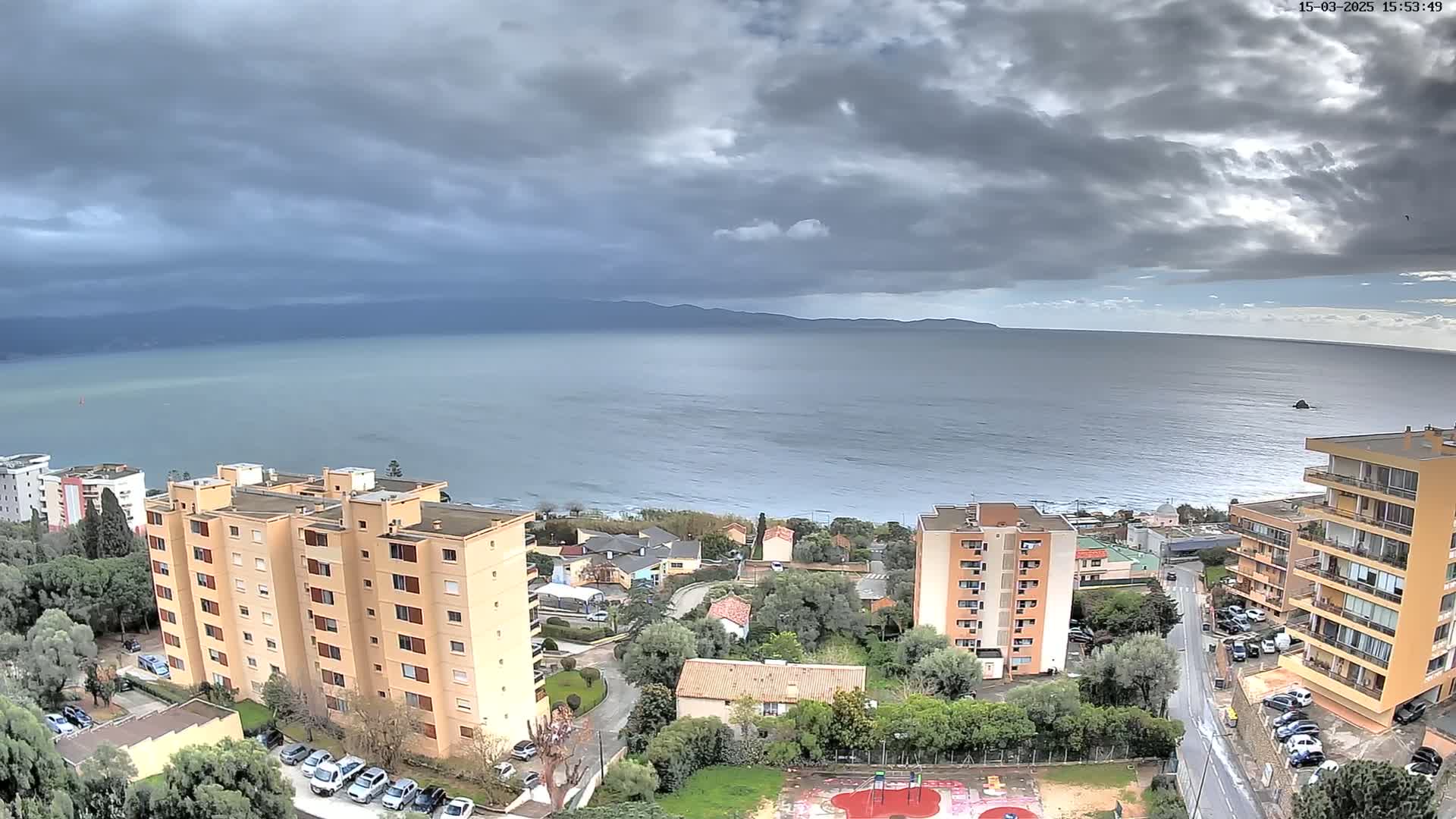A coastal view shows several multi-story buildings under a cloudy sky overlooking a calm sea and distant mountains.