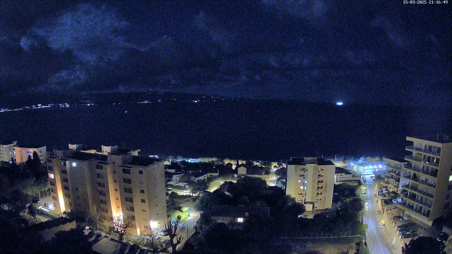 A nighttime aerial view of apartment buildings and a roadway overlooking a dark body of water under a cloudy sky.