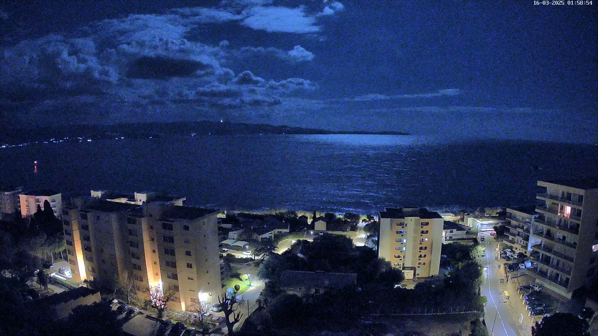 A nighttime, high-angle view shows a dark, wavy sea under a partly cloudy sky, with several multi-story buildings and streetlights illuminating a coastal town.