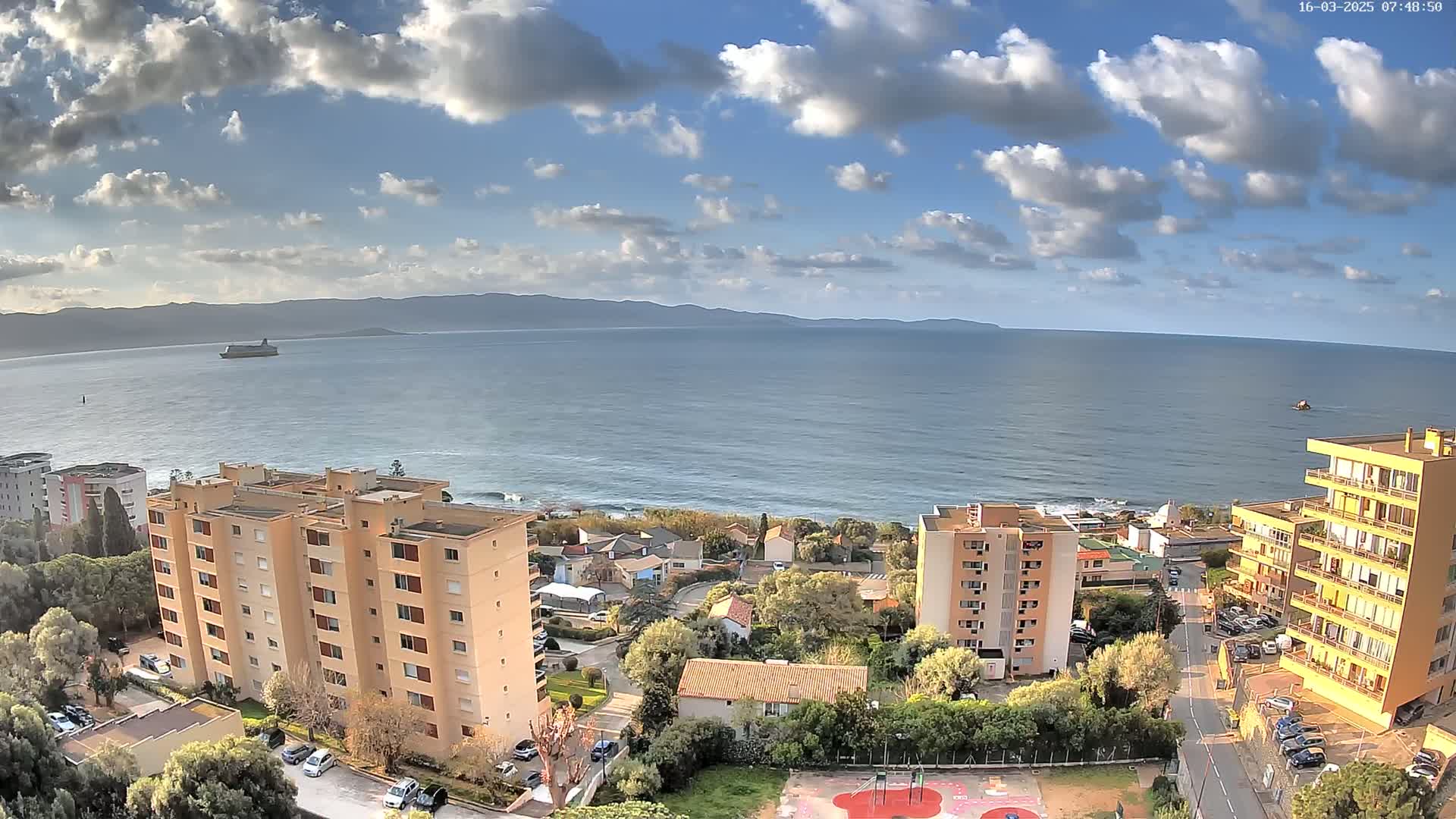 A partly cloudy day reveals a coastal scene with apartment buildings, houses, and a playground overlooking a calm sea with a large ship visible in the distance, mountains are seen on the horizon.