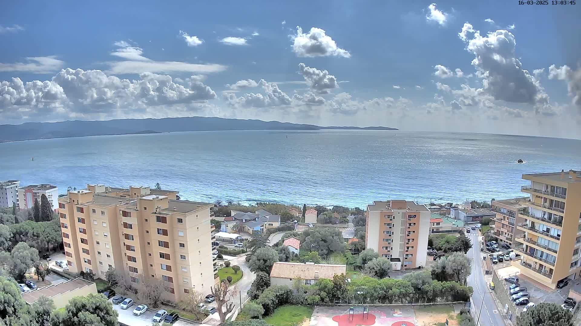 A partly cloudy day overlooks a calm ocean, with several apartment buildings and houses nestled among green trees below.