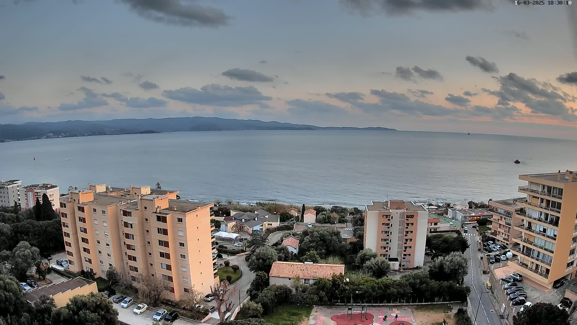 An aerial view shows a calm sea under a partly cloudy sky at dusk, overlooking coastal buildings and a playground.