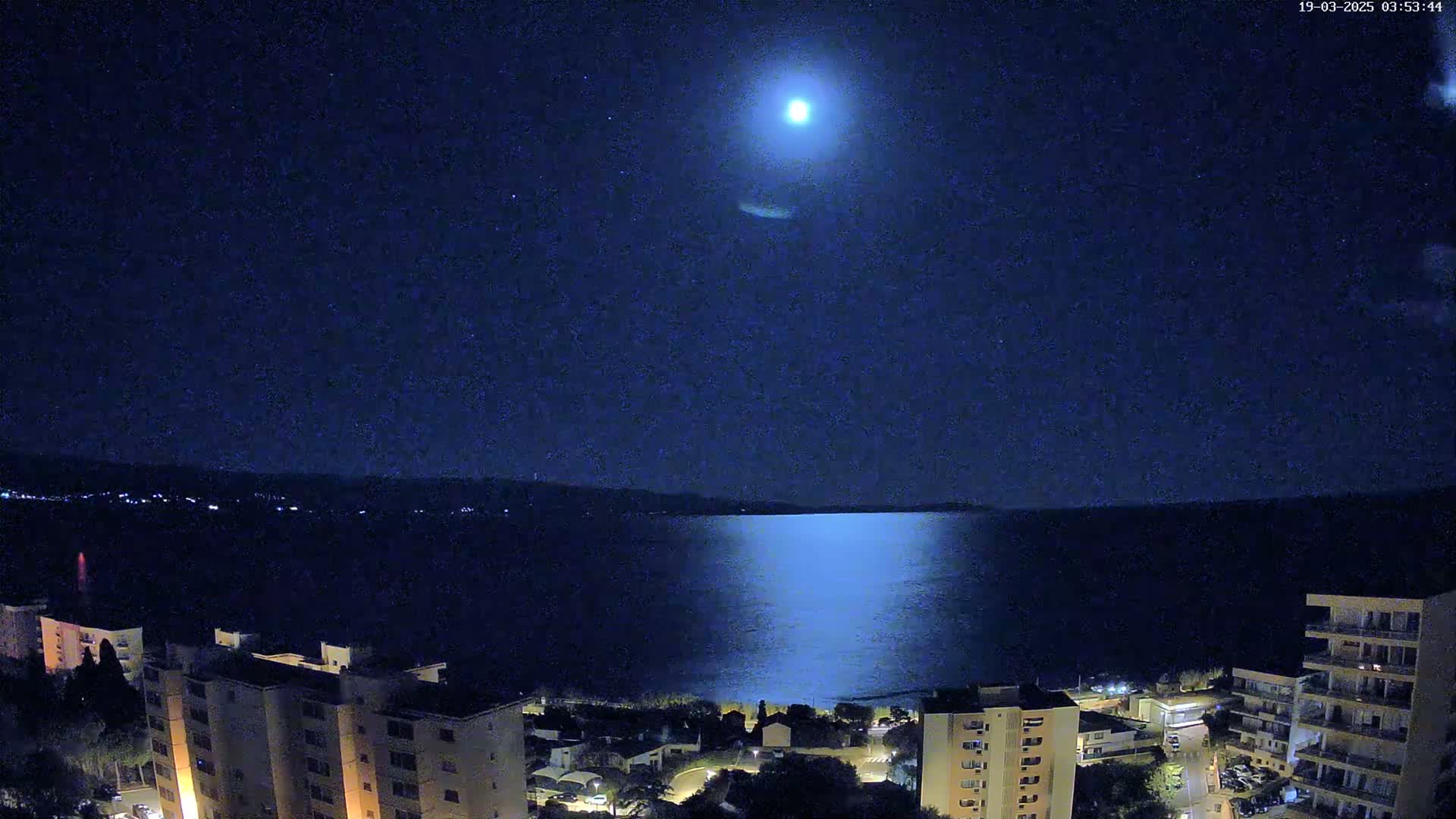 A bright moon illuminates a calm sea at night, with city lights and buildings visible along the shoreline.