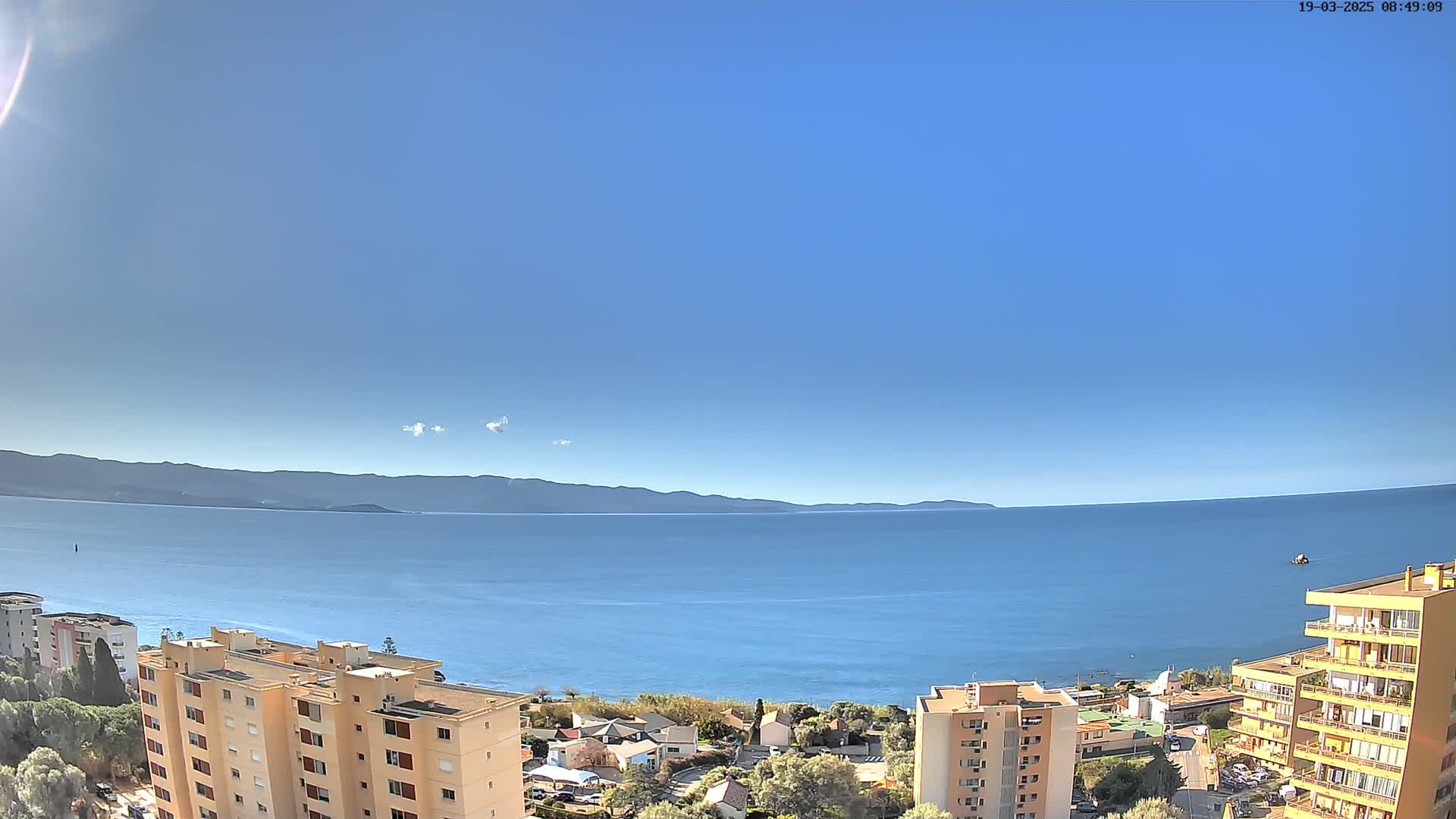 A clear, sunny day reveals a calm blue sea extending to distant mountains, with apartment buildings lining the coast.