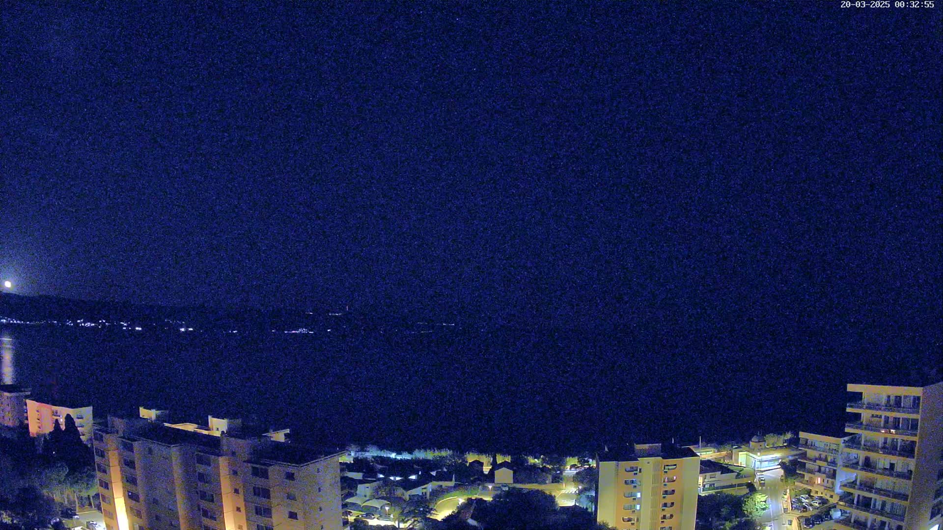 A nighttime view of a city by a body of water under a clear, dark sky shows a bright moon in the distance and city lights reflecting on the water.